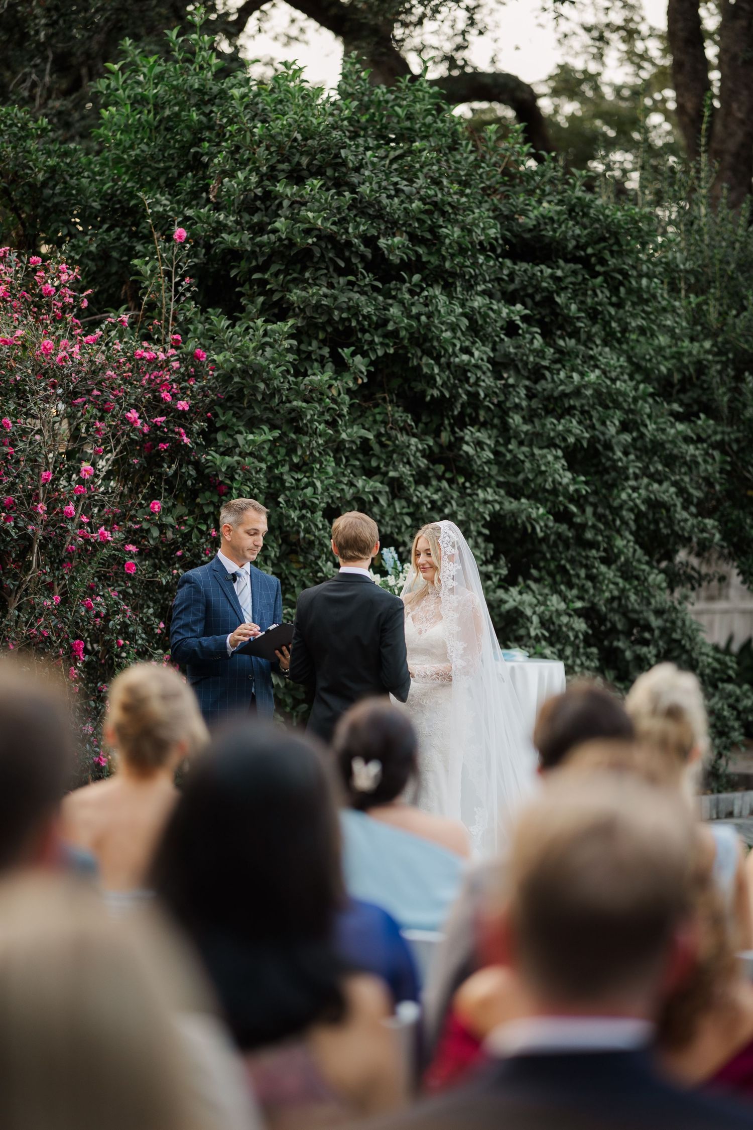 Outdoor wedding ceremony in a lush garden setting with flowering bushes and gathered guests.