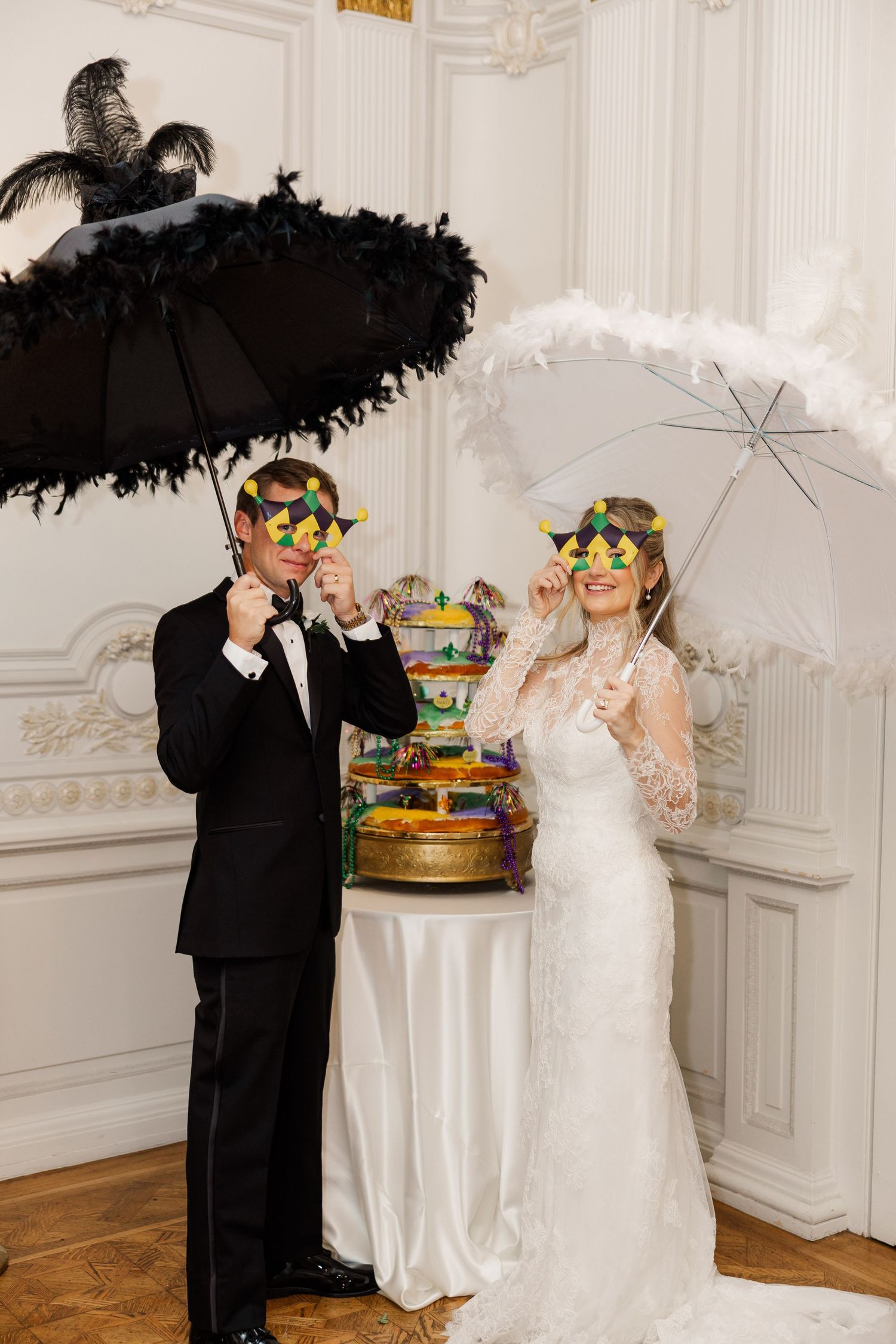 A couple in wedding attire poses with parasols and masquerade masks next to a colorful tiered cake.