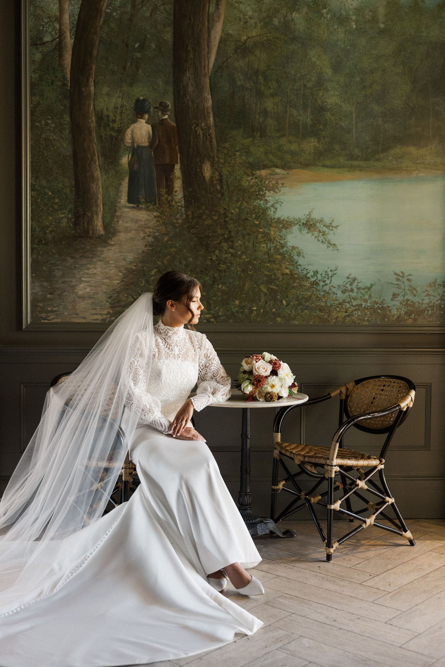 A bride in an elegant white gown sits on a porch with vintage chairs and nature-themed wall art.