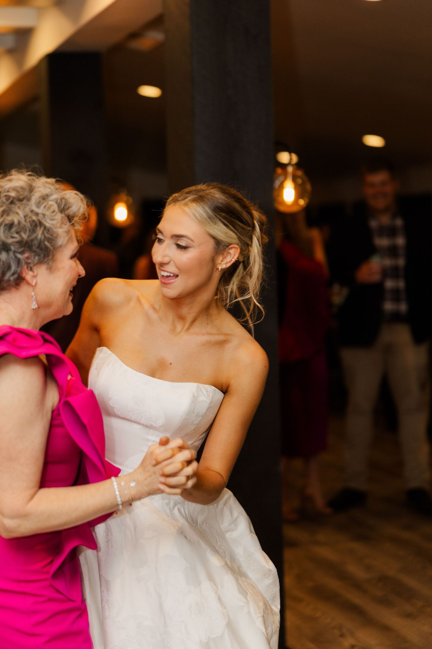 A bride in a strapless gown shares a dance with a guest in a bright pink dress.