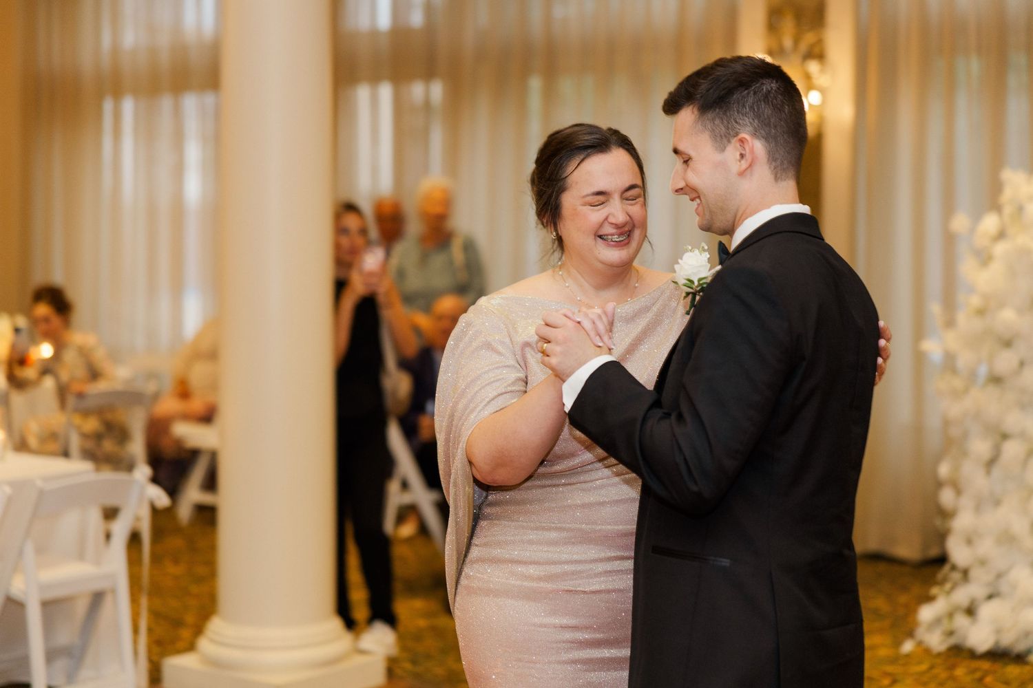 Wedding couple sharing a joyful dance moment in an elegant ballroom with columns and warm lighting.