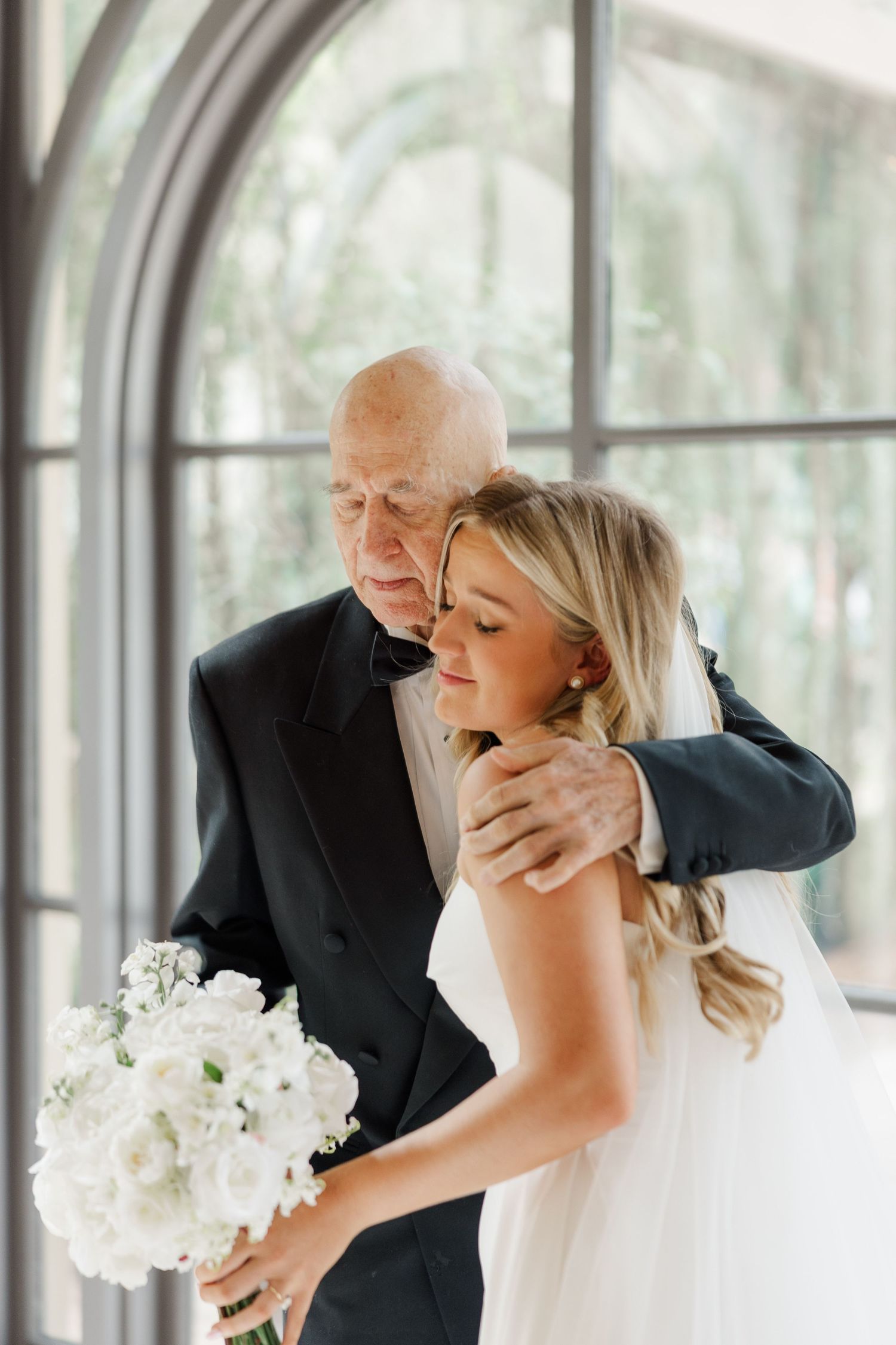 Emotional wedding first look moment in a bright sunlit room with white floral bouquet.
