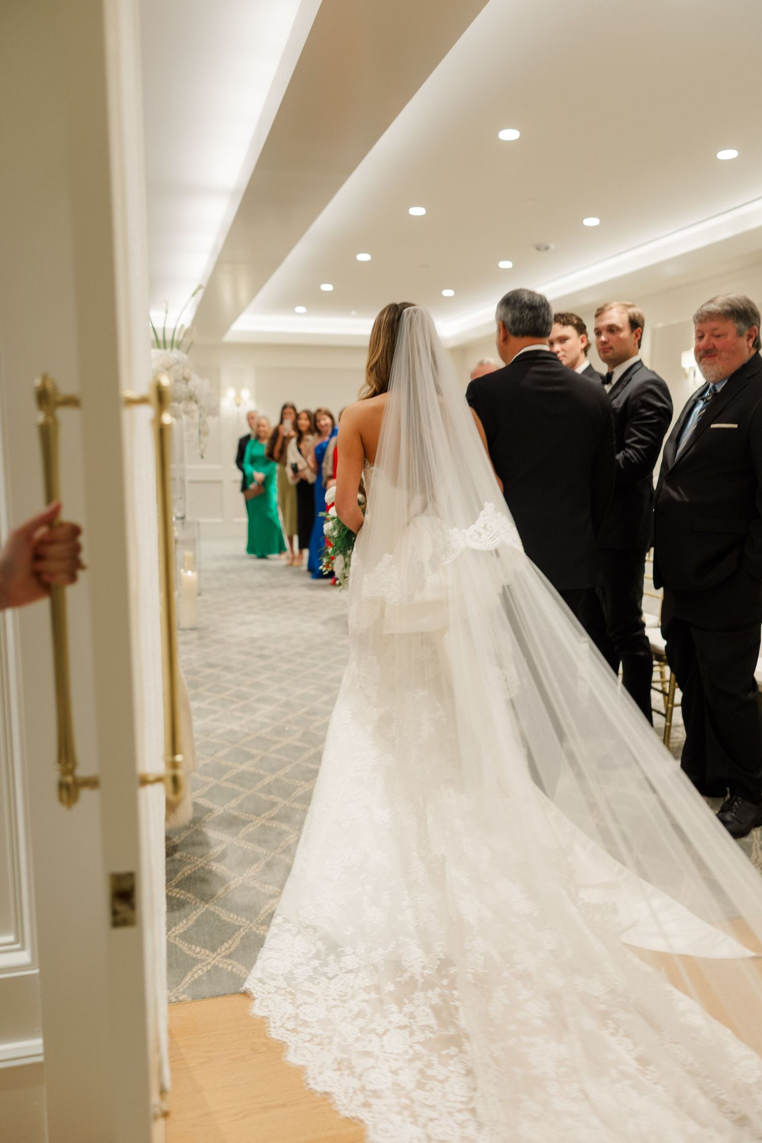 Cathedral length lace veil trailing behind bride in elegant white hallway with recessed lighting.