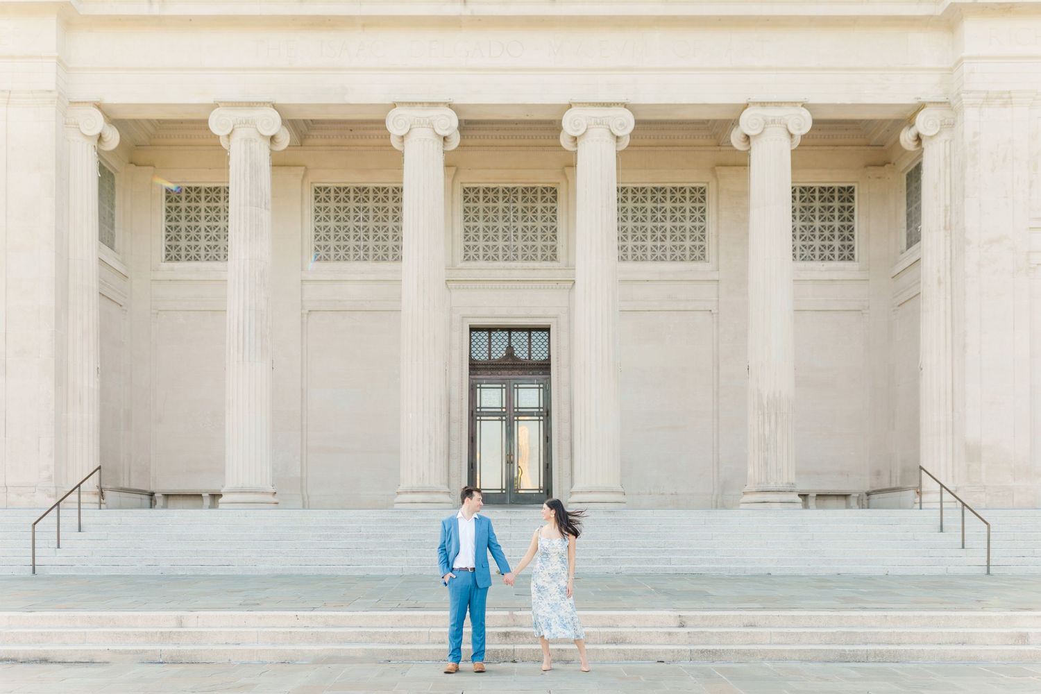 A couple in blue suit and white dress stands on marble steps in front of a grand neoclassical building with columns.