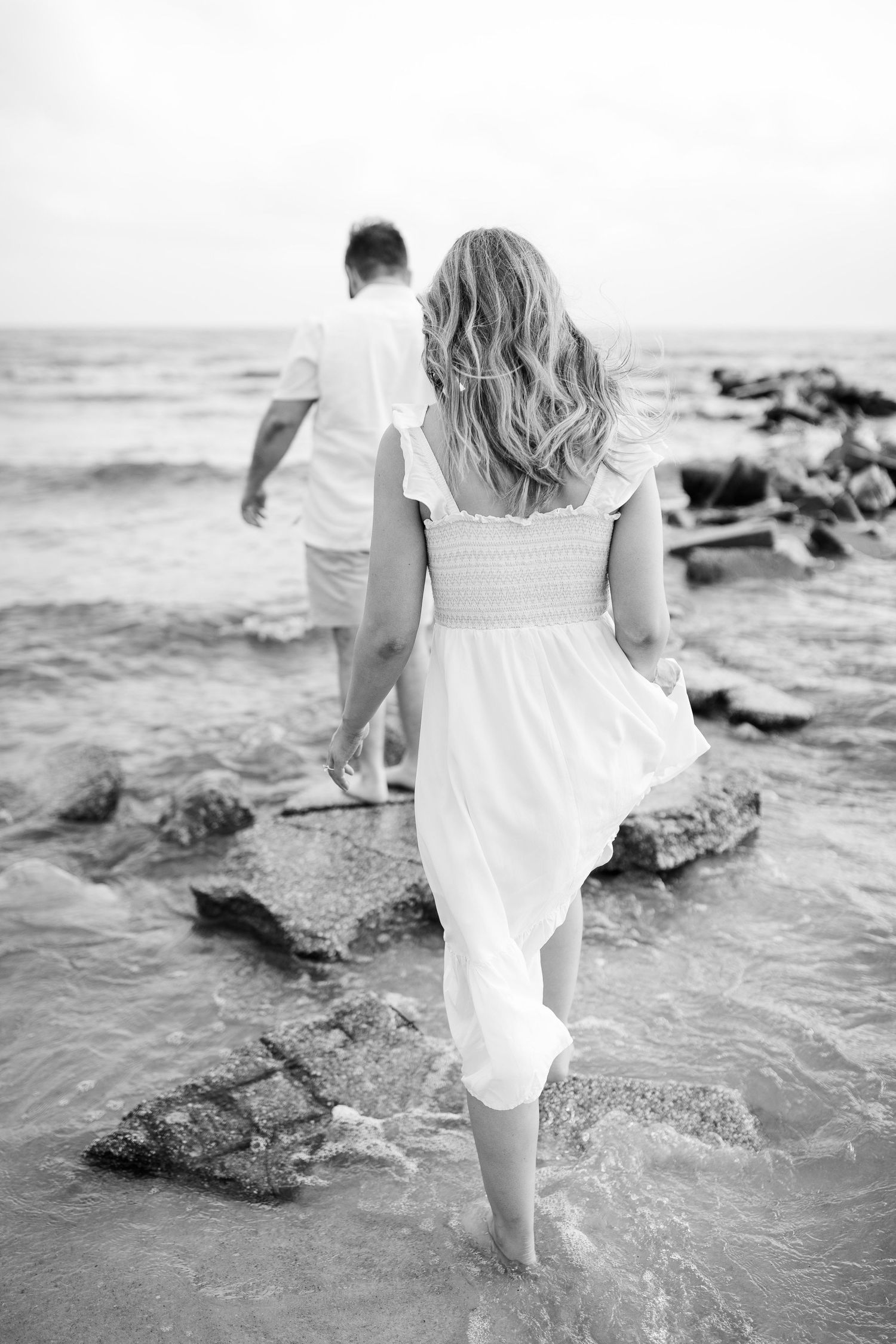 A black and white photo of a couple walking on rocky coastal shores with waves crashing nearby.