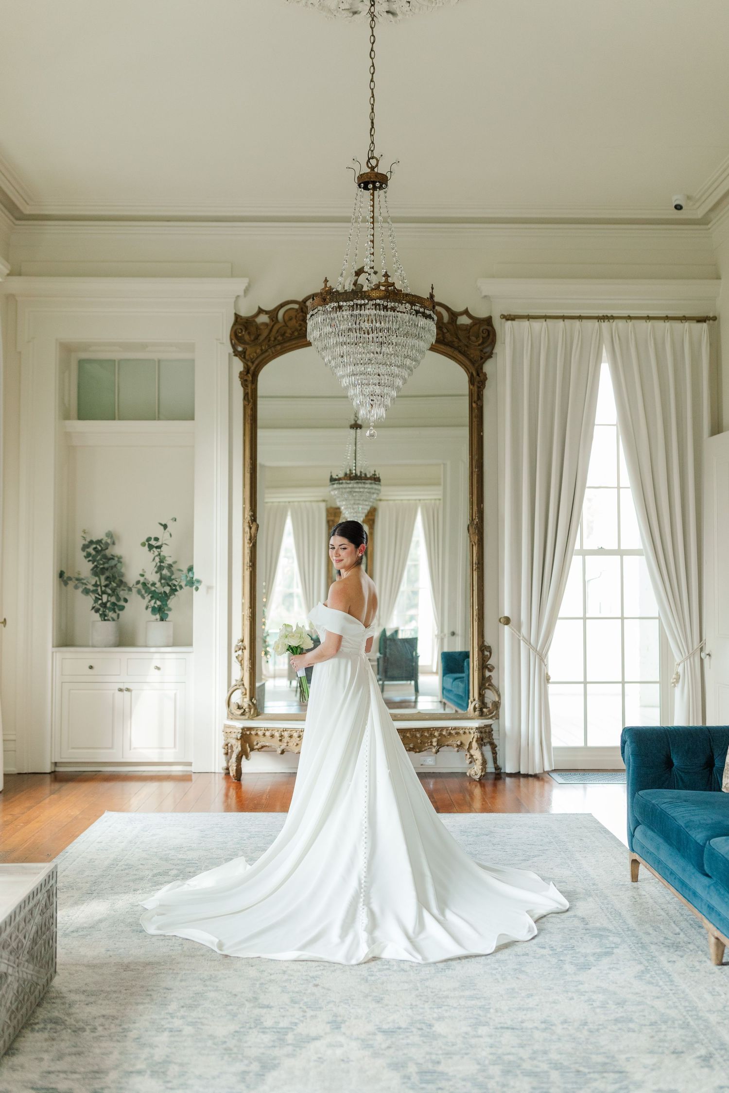 A bride in an elegant white wedding gown poses in front of an ornate mirror in a luxurious room with crystal chandelier.