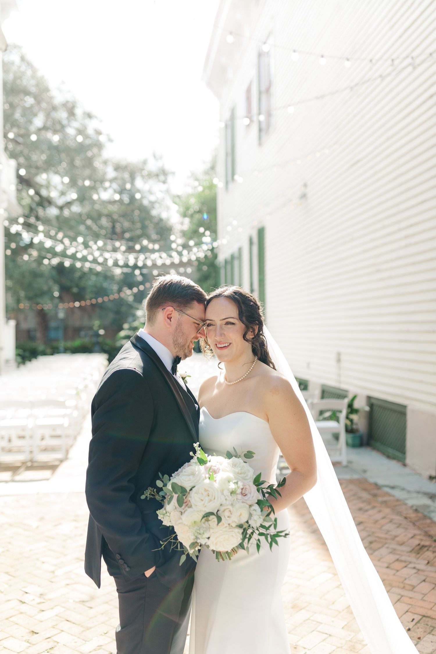 A romantic wedding moment captured in soft sunlight with string lights and a white brick building in the background.