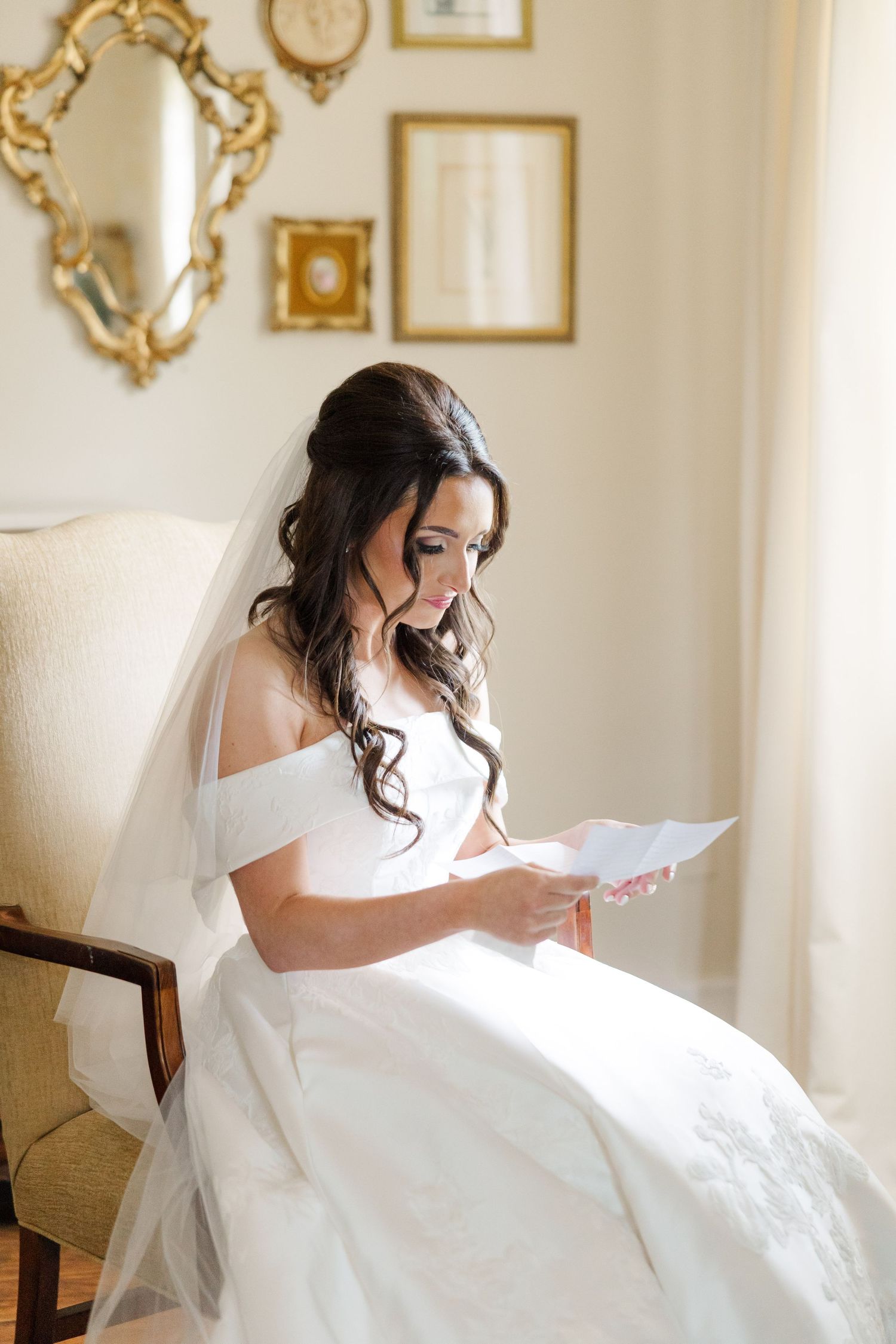 Bride in white gown reads a letter while sitting in an elegant room with gold-framed mirrors.