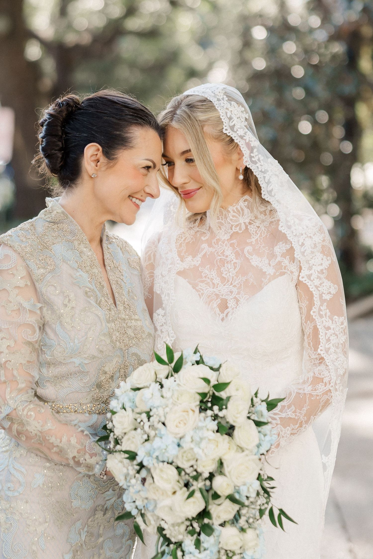 Two people in elegant lace wedding attire share an intimate moment while holding a white floral bouquet.