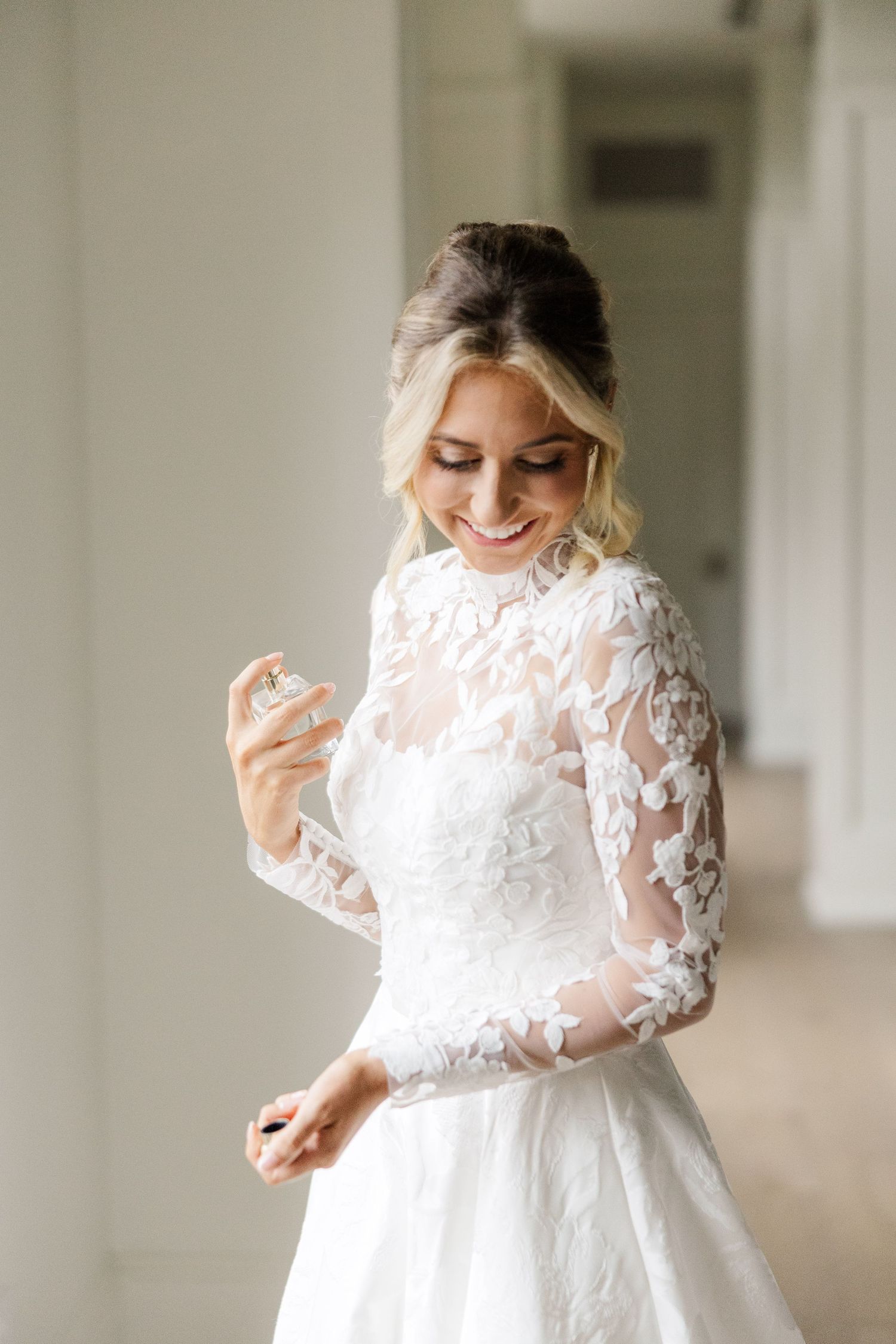 A bride in a long-sleeved lace wedding gown twirls and smiles in a bright indoor setting.