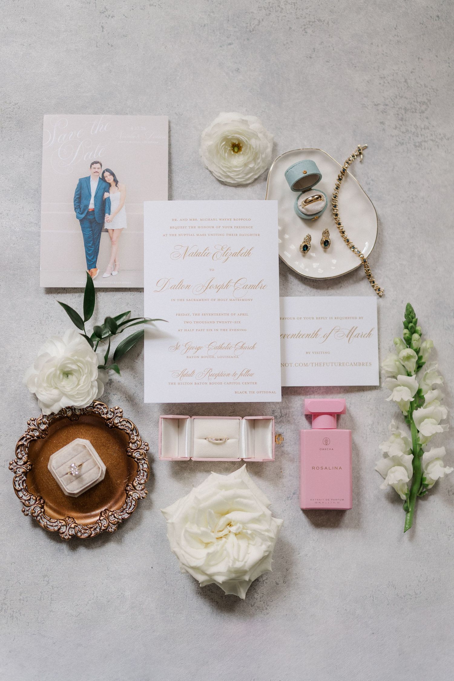 Elegant wedding flat lay featuring gold script invitations, white ranunculus, macarons, ring box, and pink perfume on gray background.