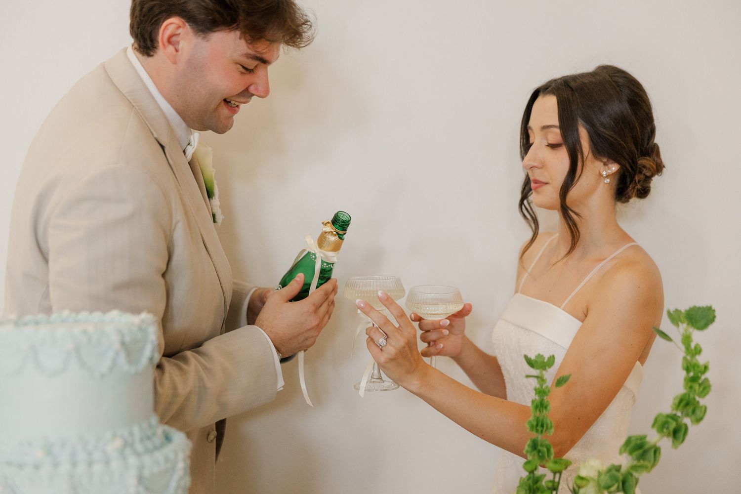 Groom in tan suit opens champagne bottle while bride in white dress holds crystal flutes at wedding reception.