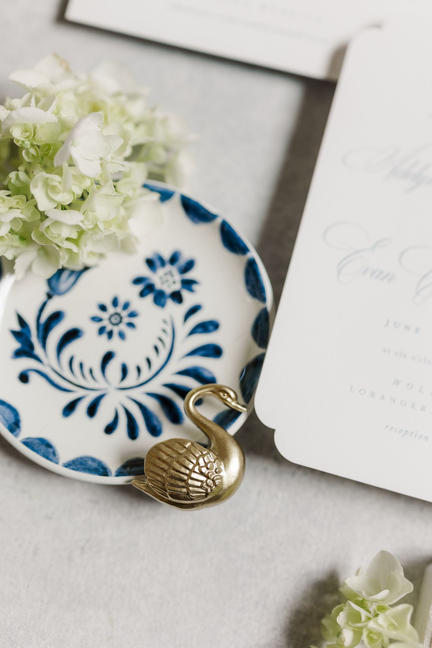 Blue and white floral ceramic dish with gold swan figurine beside wedding invitation and white hydrangeas.