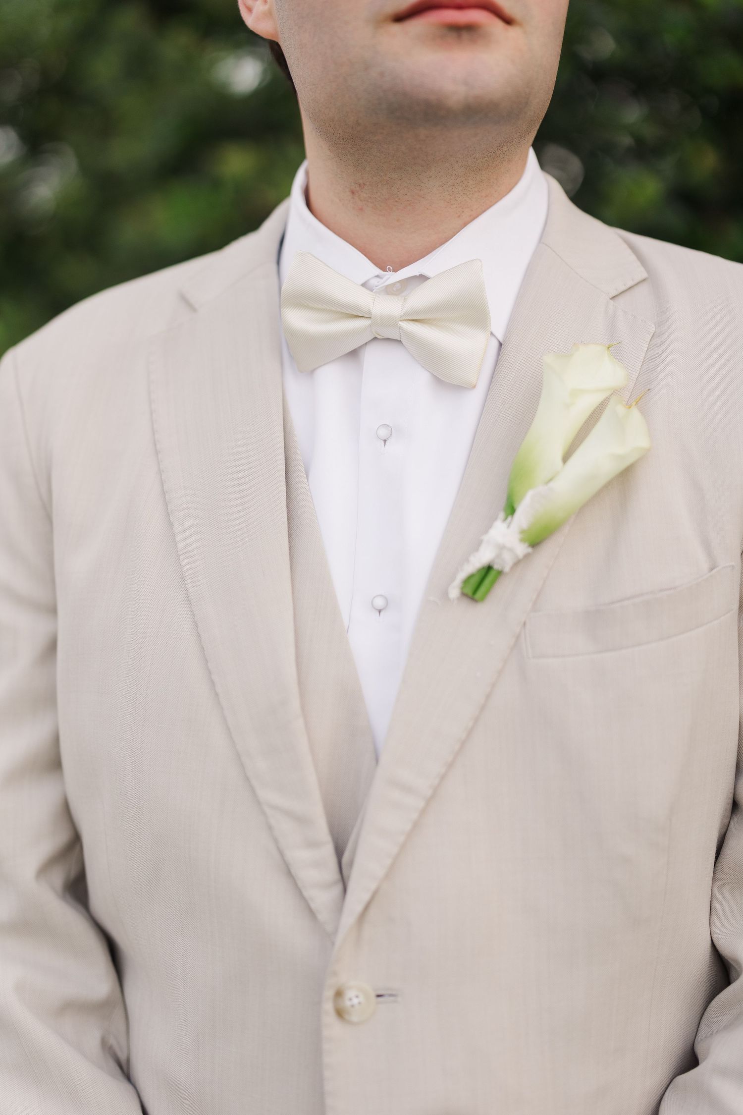 Groom in beige suit with bow tie and white calla lily boutonniere at an outdoor wedding ceremony.
