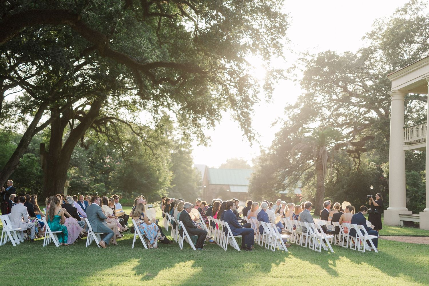 Outdoor wedding ceremony on sunlit lawn with white chairs, large oak trees, and a white columned estate backdrop.