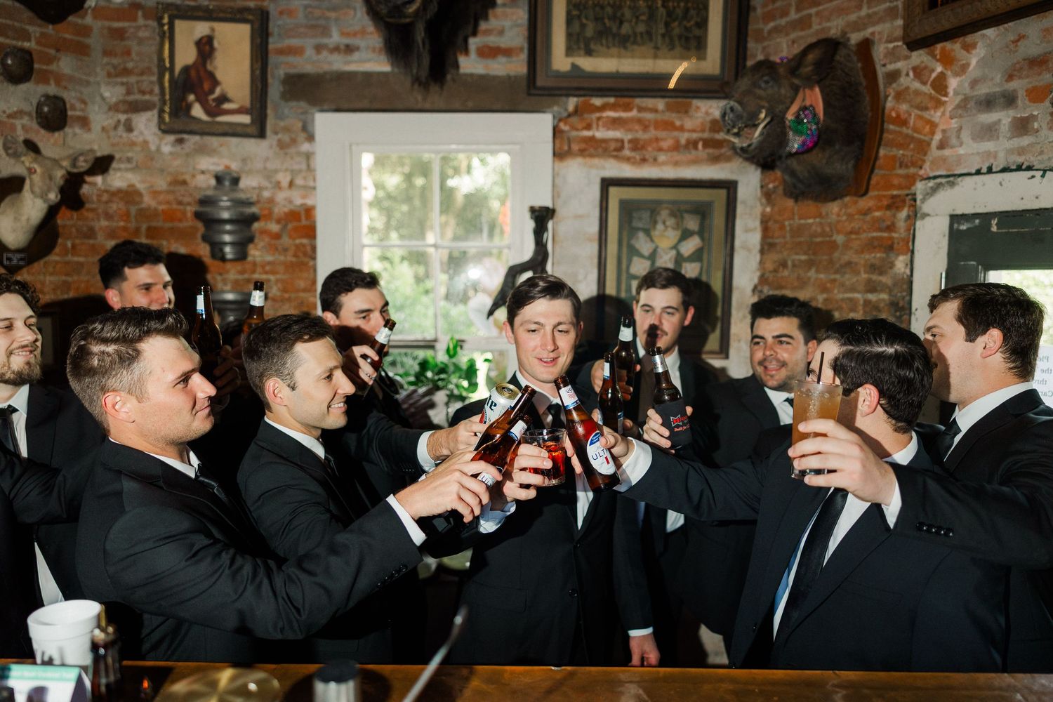 Groomsmen in black tuxedos toasting with beer bottles at a rustic bar during wedding celebrations.