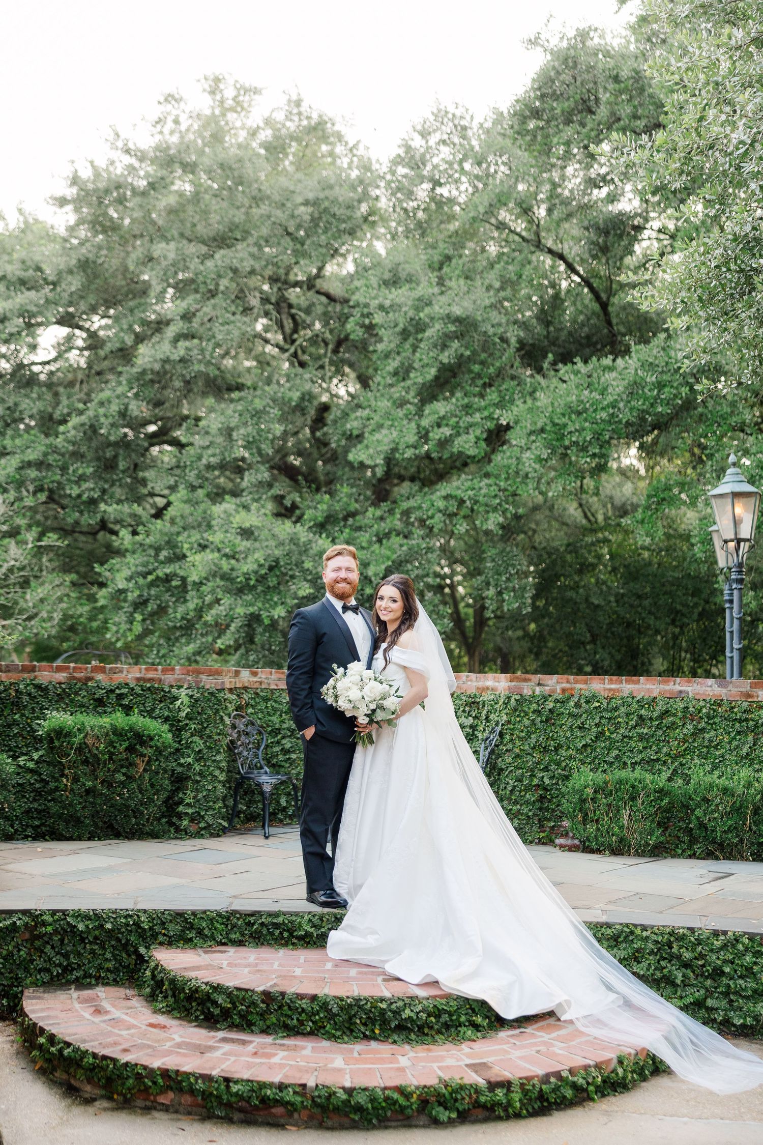 A romantic wedding couple poses in a garden setting with lush greenery and a curved brick path.
