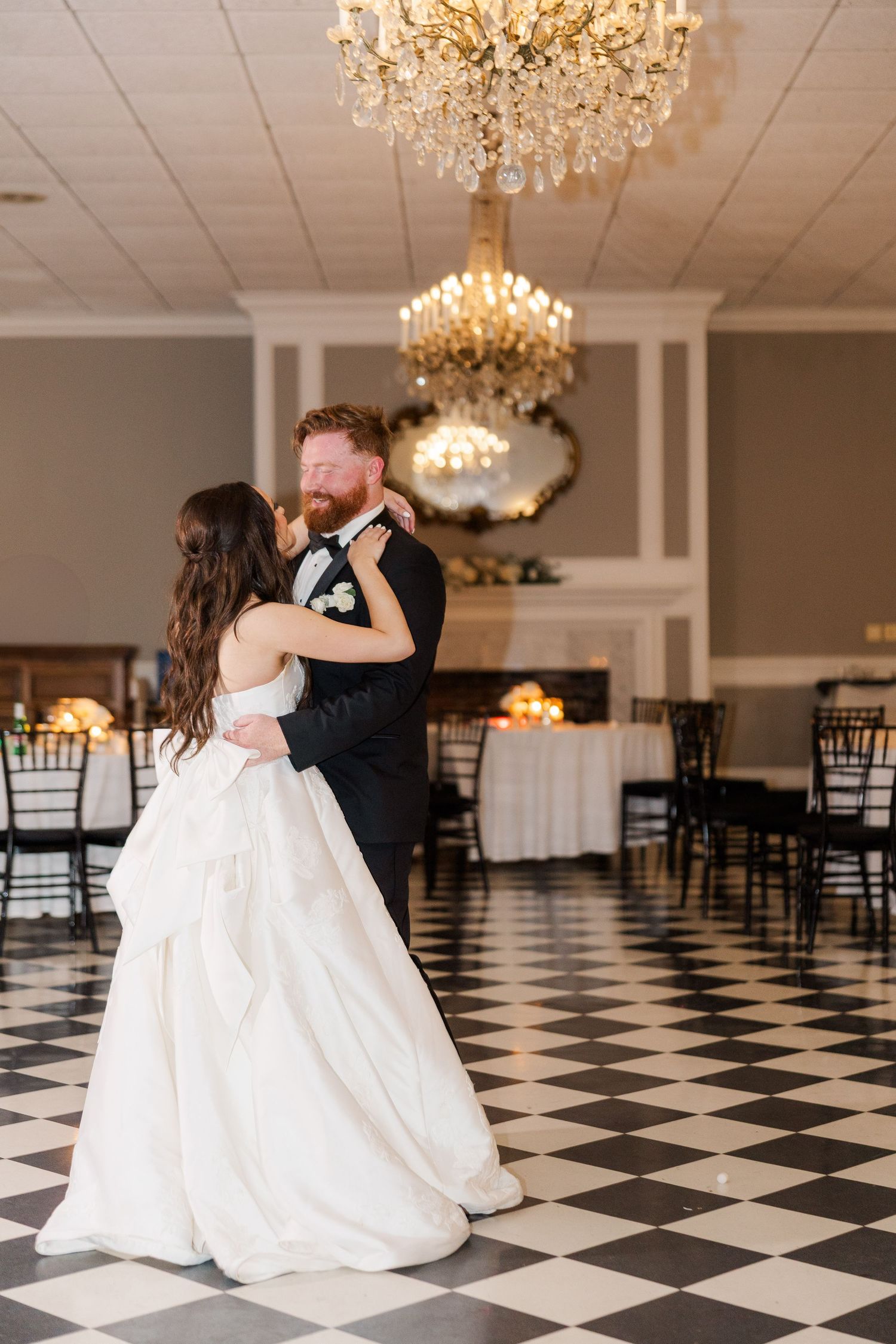 Couple shares first dance under crystal chandeliers on black and white checkered floor.