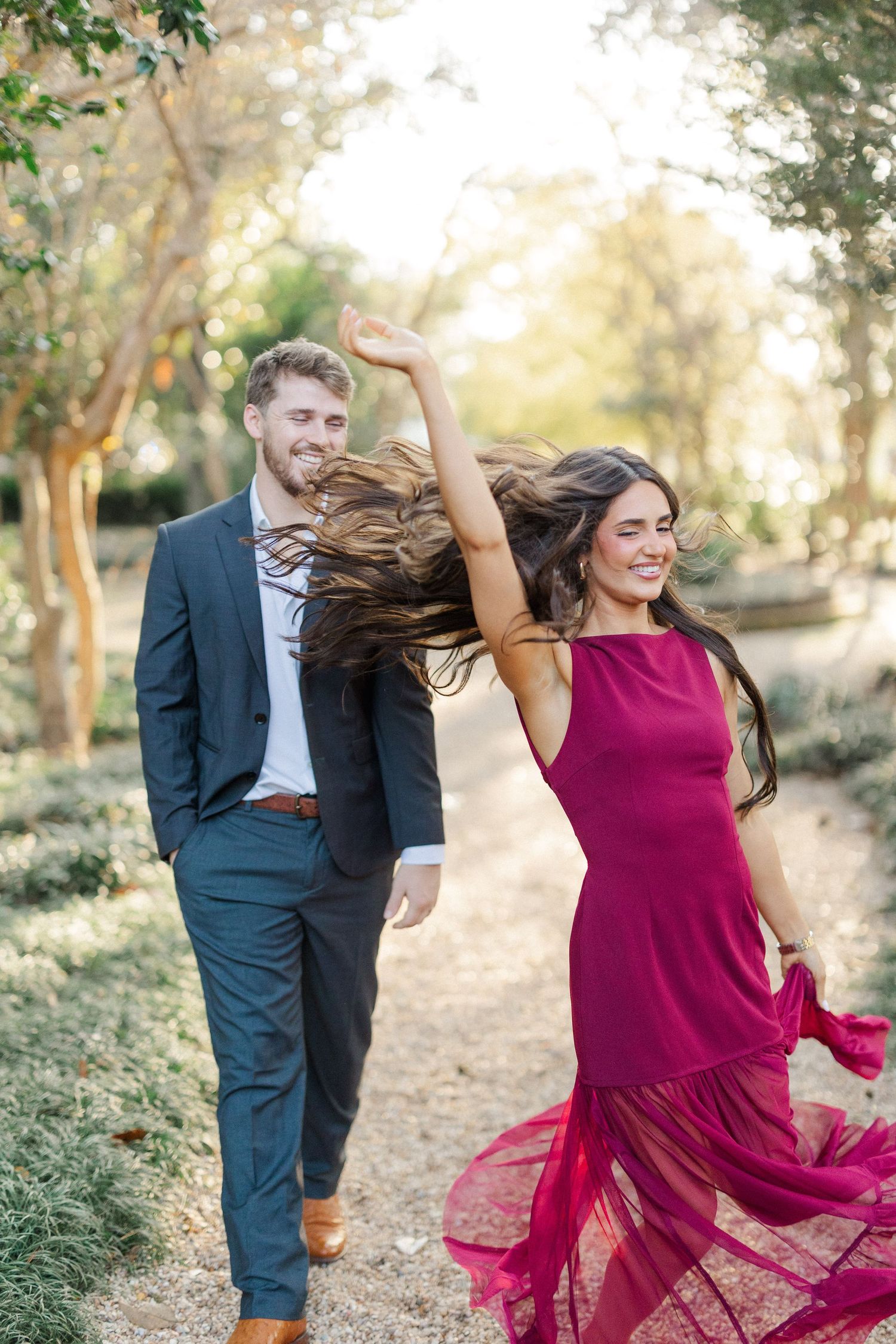 A couple dances joyfully in a garden as her burgundy dress flows in the wind.
