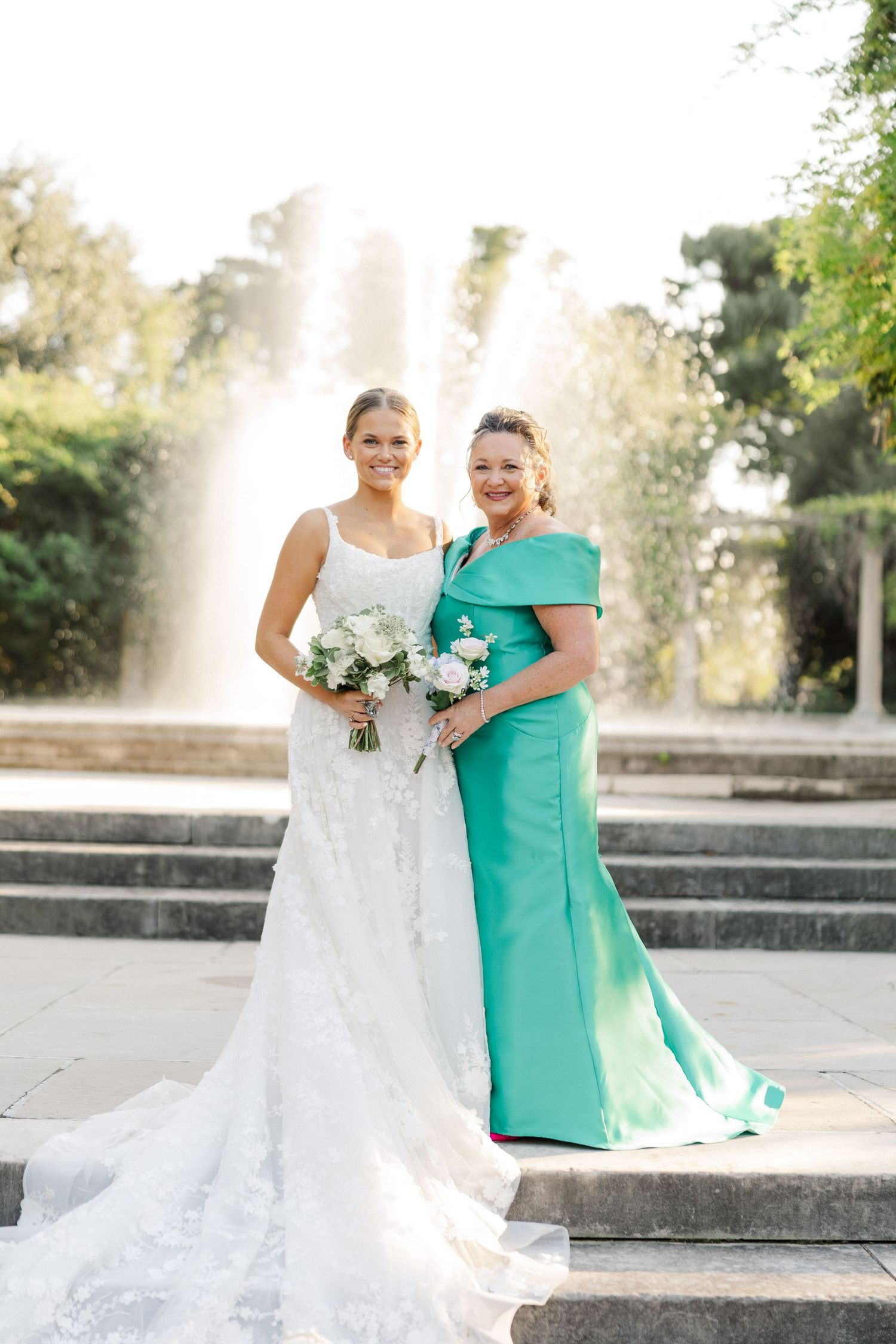 Elegant wedding portraits on stone steps with a white gown and emerald green dress against a fountain backdrop.
