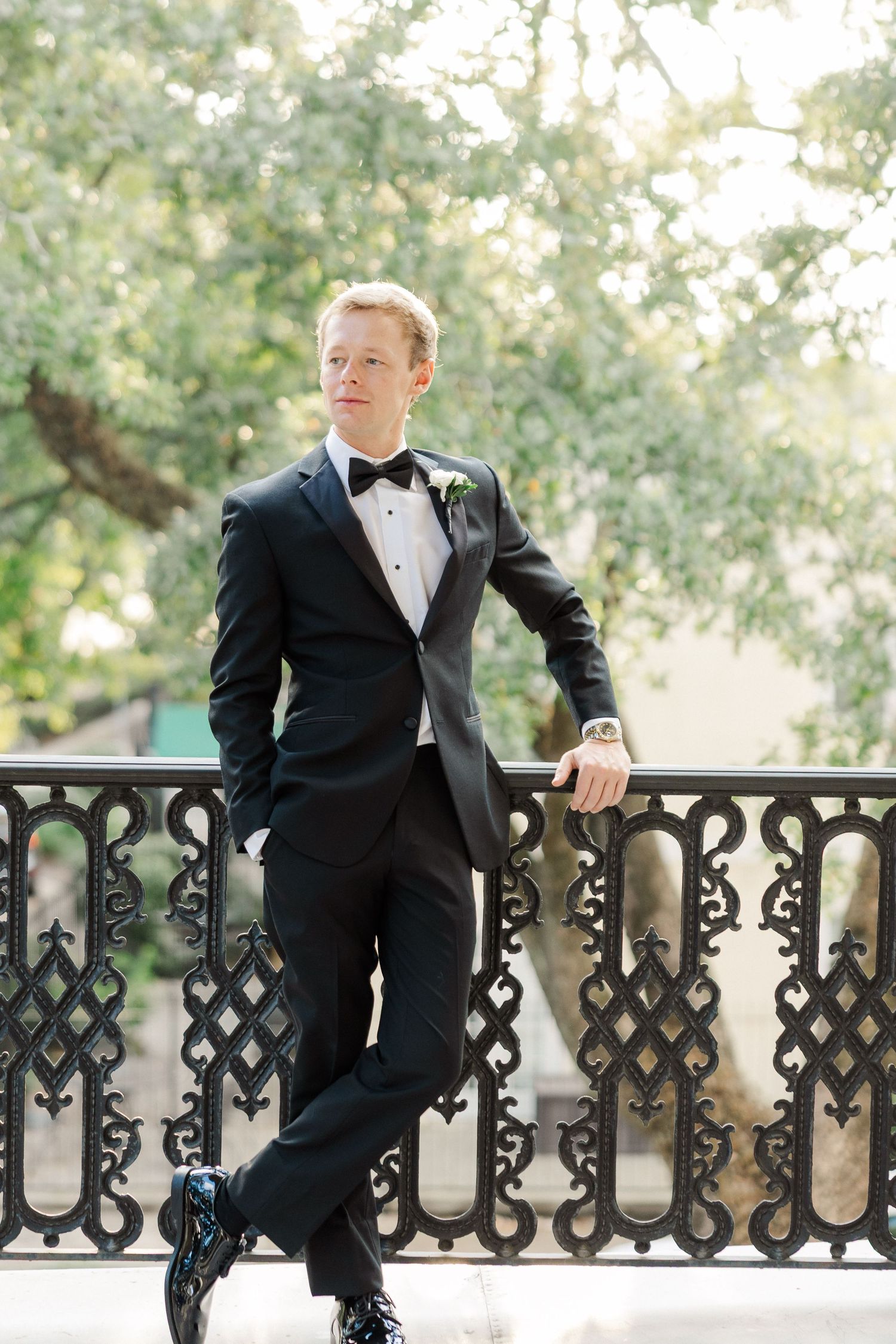 A groom in a black tuxedo poses on an ornate balcony railing with decorative ironwork patterns.