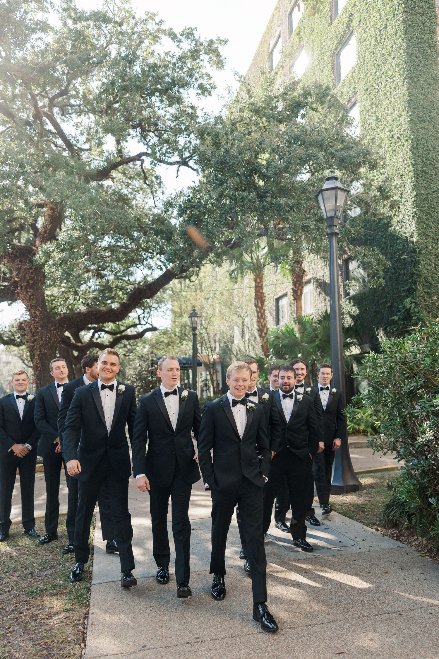Group of groomsmen in black tuxedos walking along tree-lined pathway.