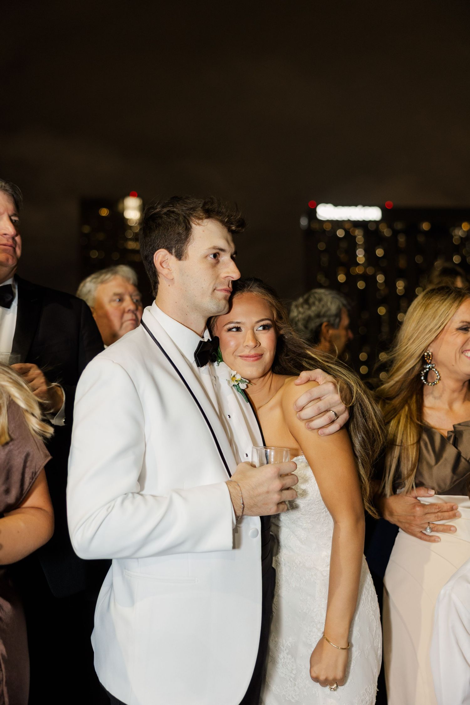 Wedding guests dance and celebrate at an evening reception with city lights in background.