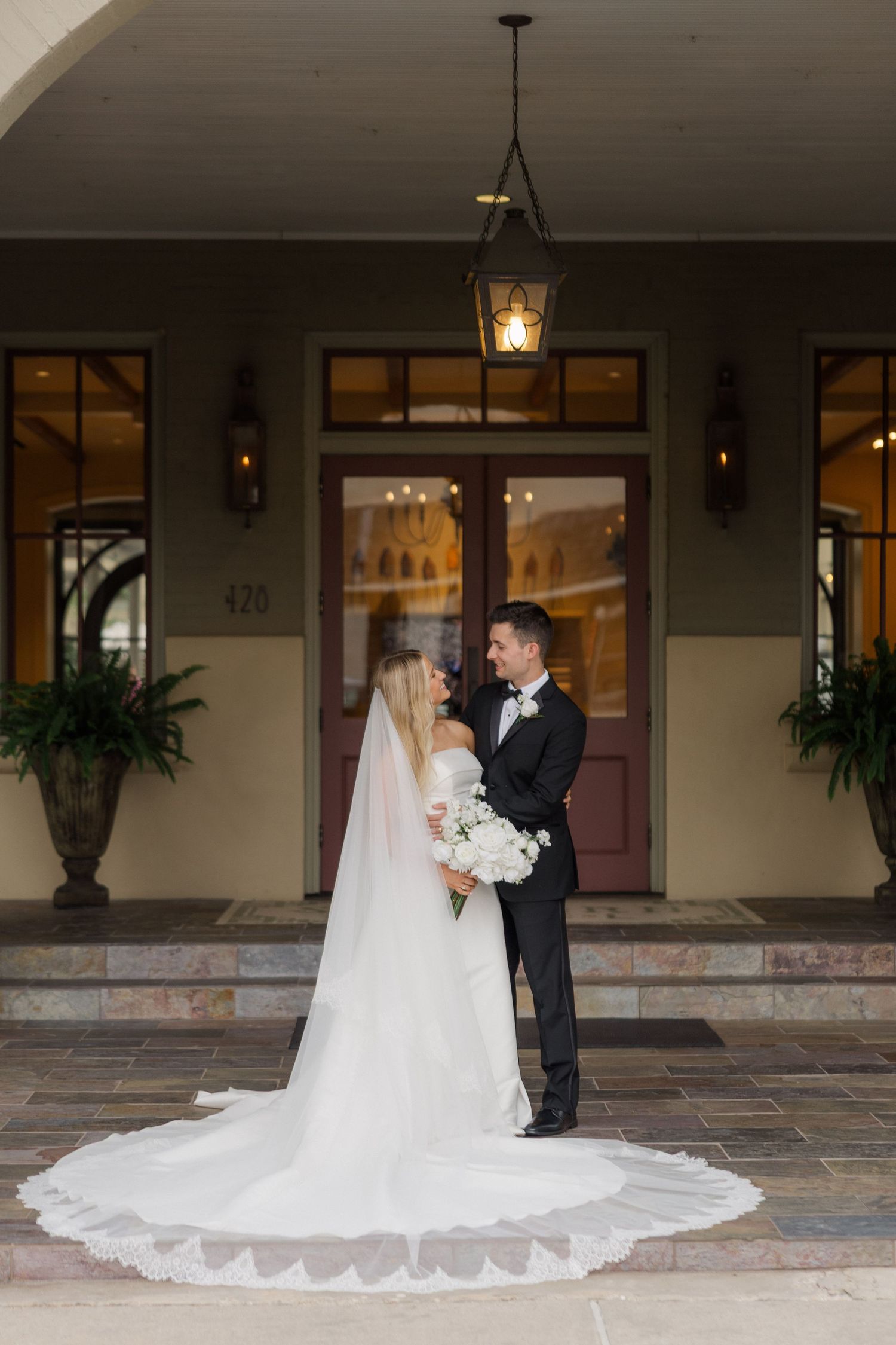 Elegant wedding couple poses under covered walkway with beautiful architectural details and hanging lanterns.
