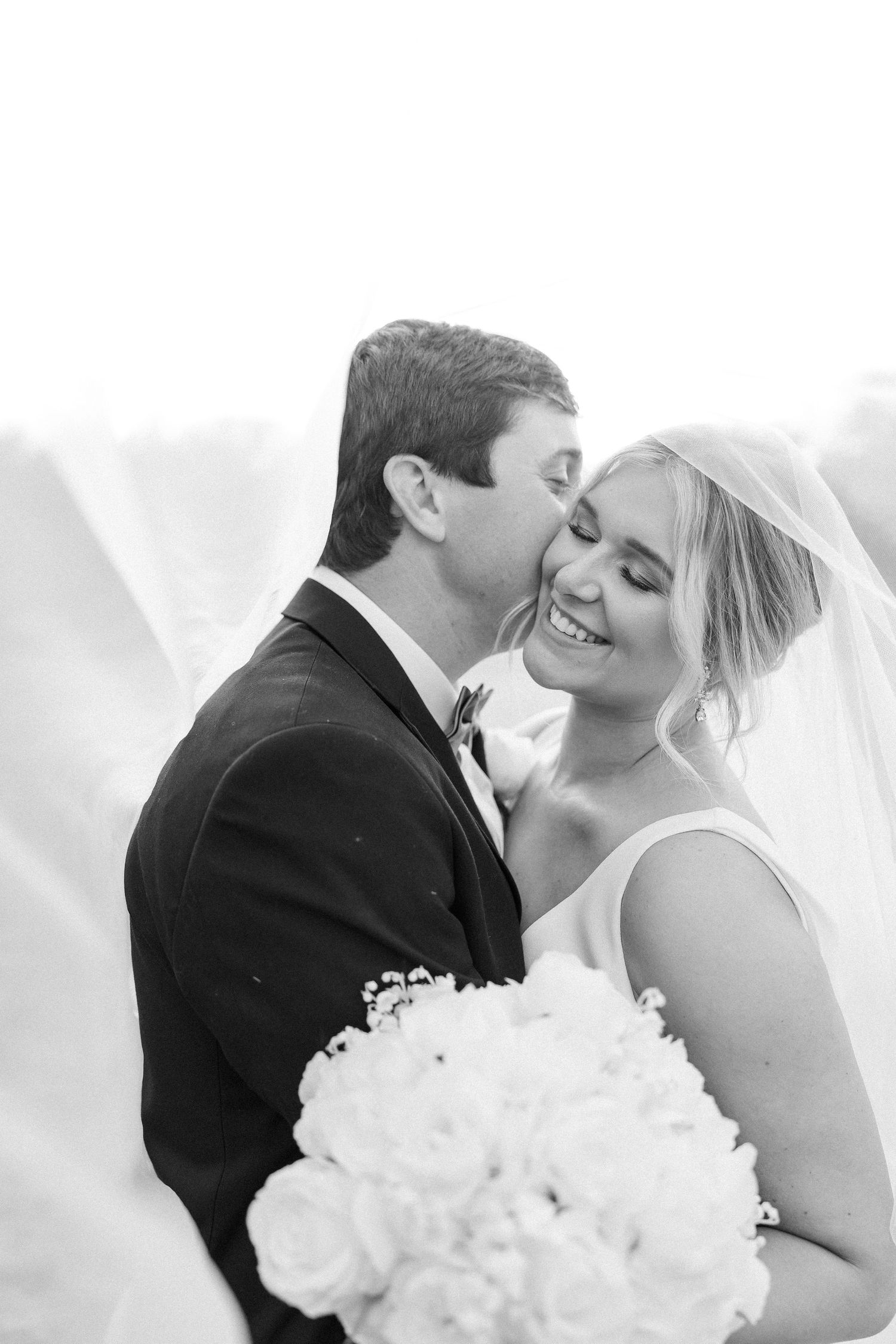 Black and white wedding photo of smiling bride holding rose bouquet as groom kisses her cheek beneath flowing veil.