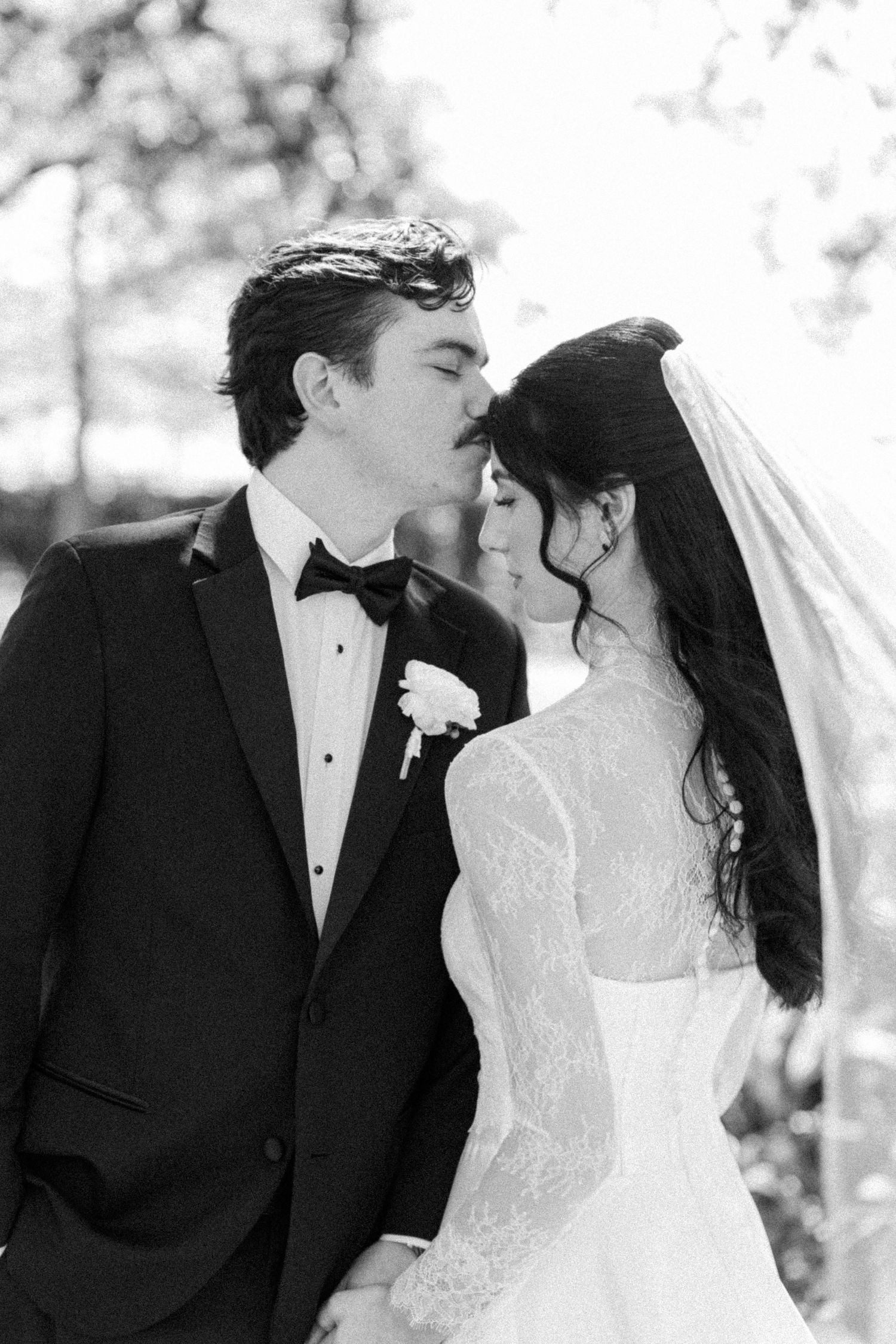 Black and white portrait of groom kissing bride wearing lace dress and veil outdoors.