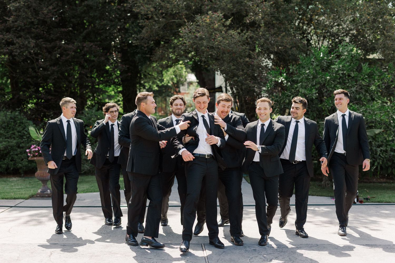 Groomsmen in black suits walking outdoors, laughing and celebrating together on a sunny wedding day.