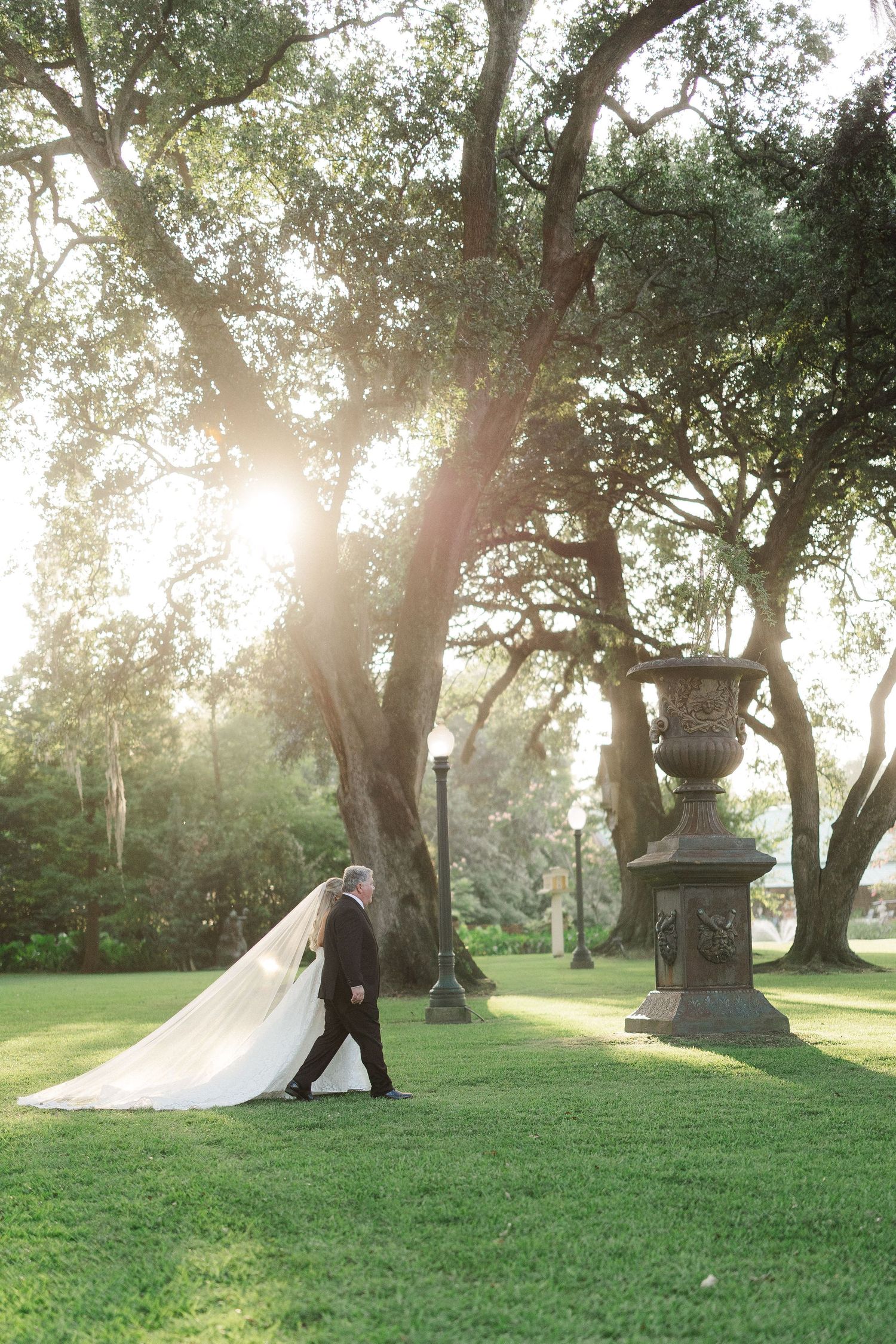 Bride and groom stroll across sunlit green lawn with long cathedral veil trailing among grand oak trees.