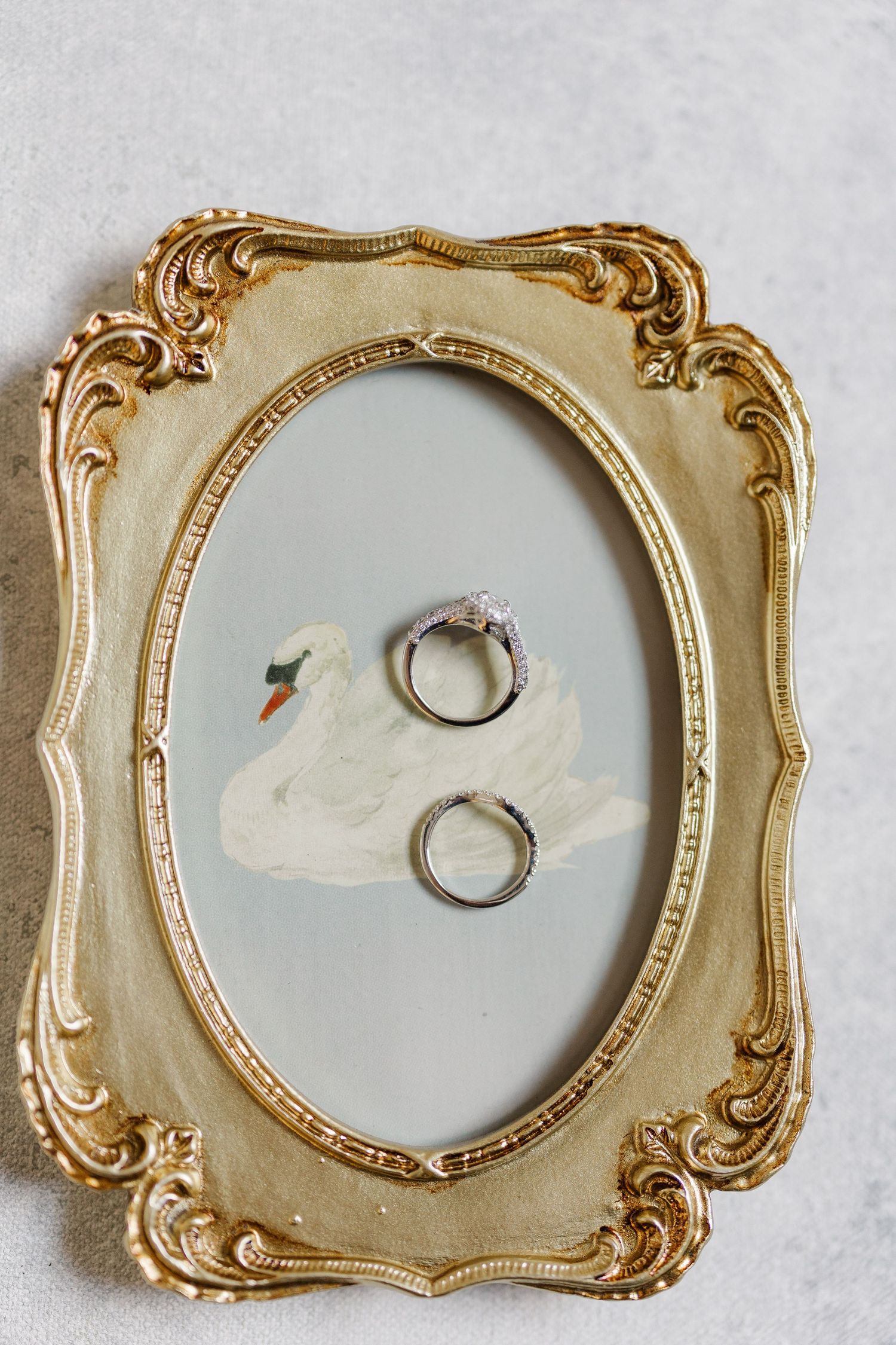 Two wedding rings displayed on a vintage gold ornate mirror frame with a painted swan illustration.