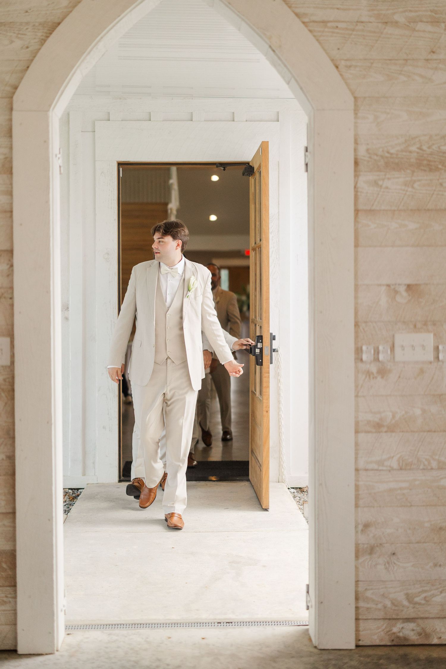Dapper groom in beige suit walking through arched doorway of rustic wedding venue.