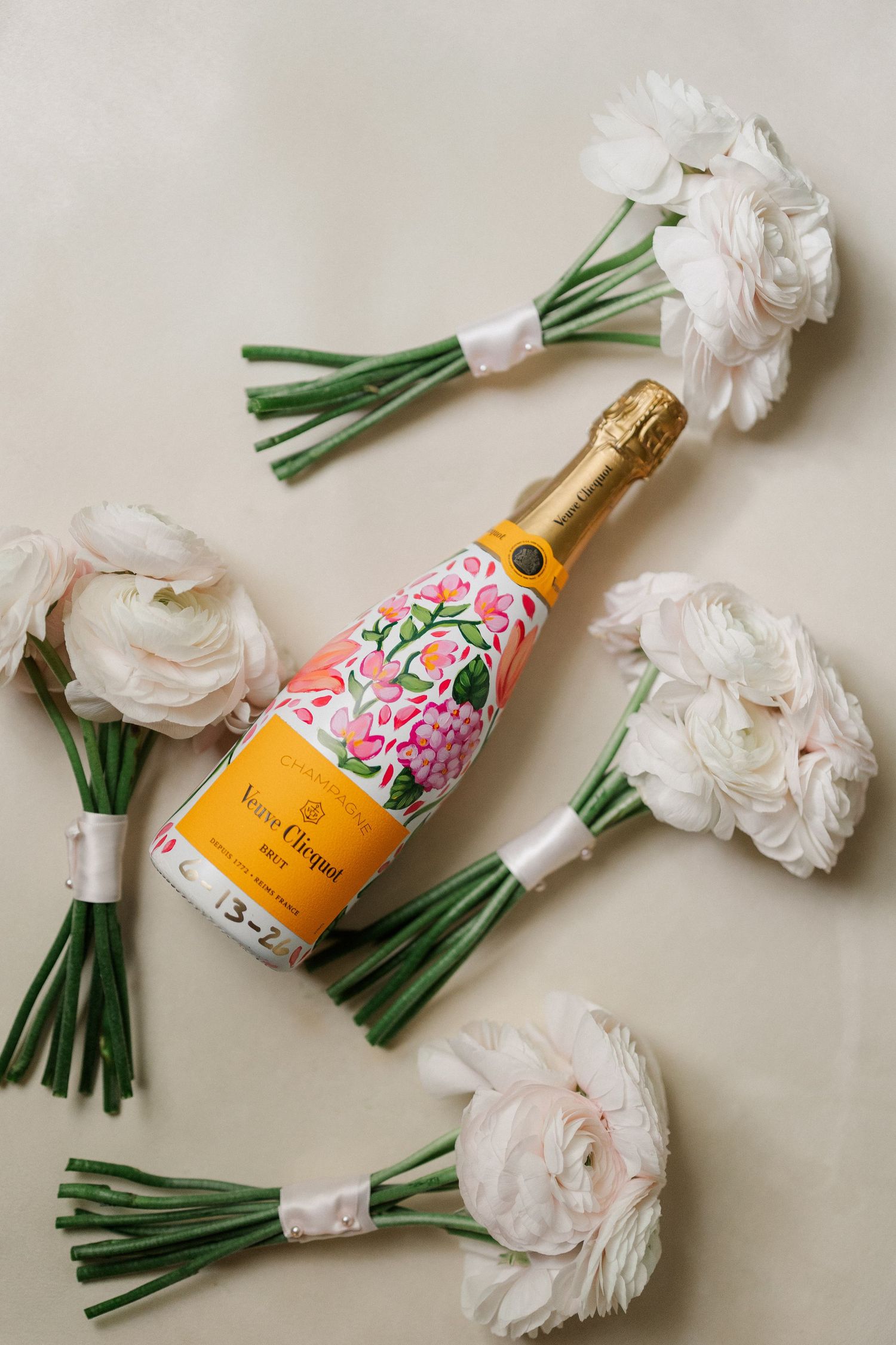 Veuve Clicquot champagne bottle with floral label surrounded by white ranunculus bouquets on beige background.