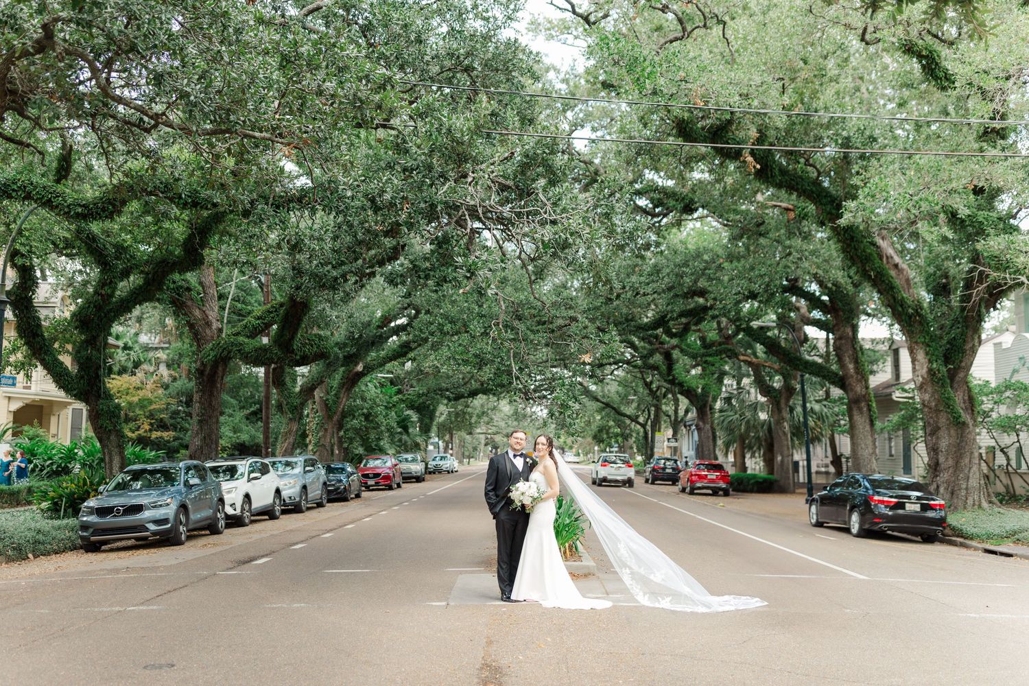 A couple embraces under a canopy of oak trees on a street lined with parked cars in New Orleans.