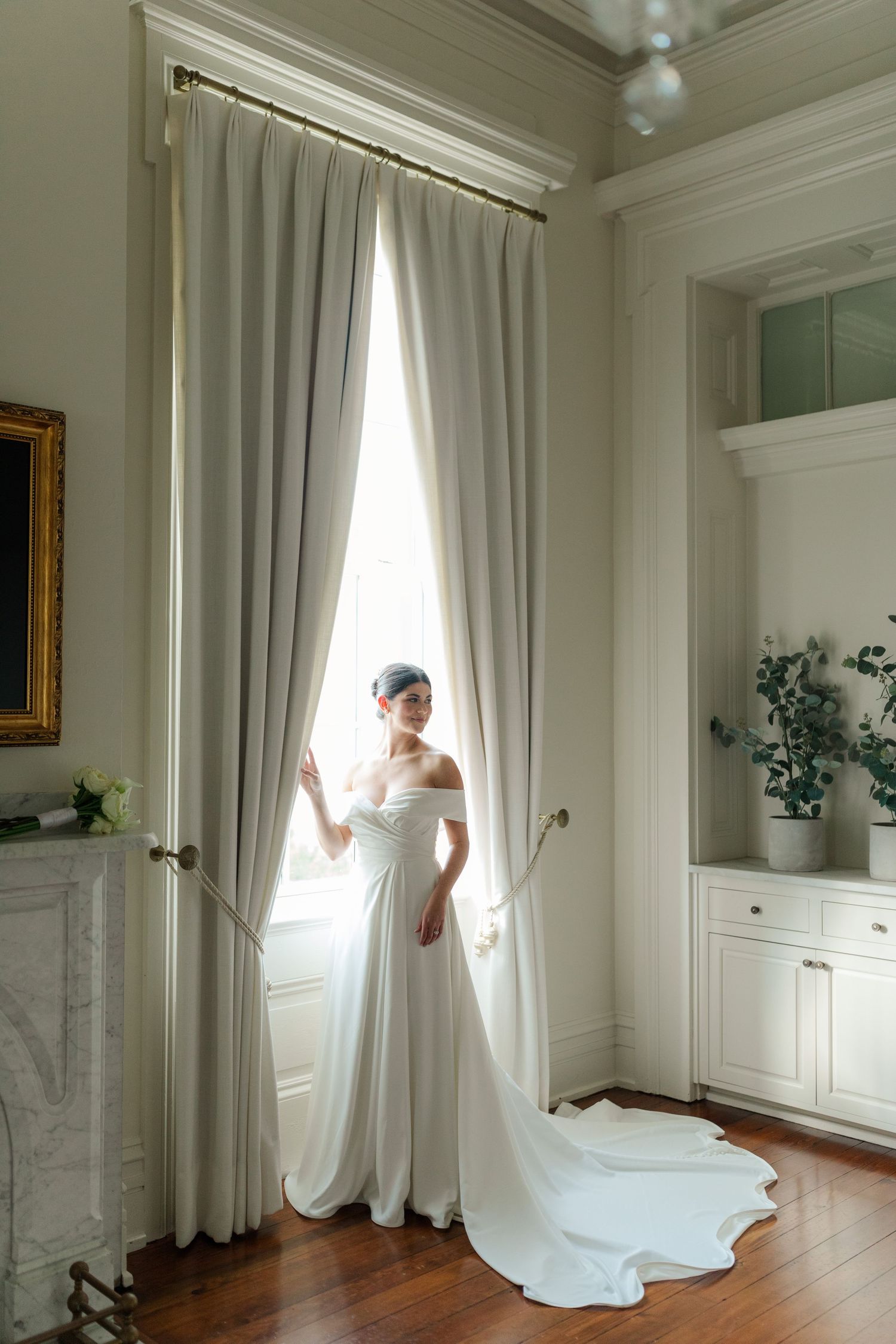 Classic bridal portrait by a window featuring an off-shoulder white gown in an elegant white room.