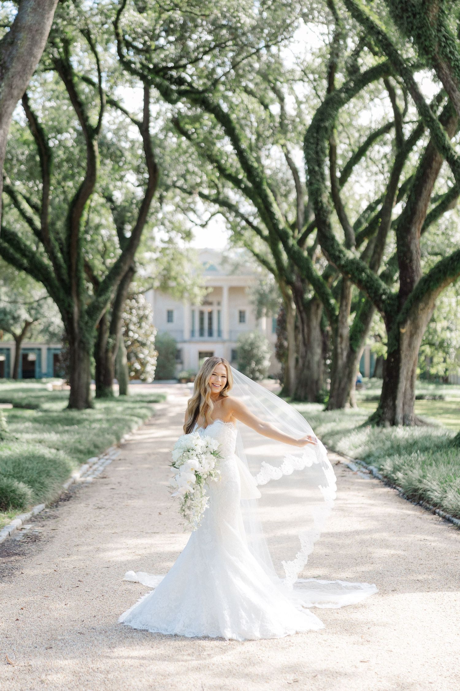 A bride in a flowing white dress twirls gracefully beneath a canopy of oak trees on a garden path.