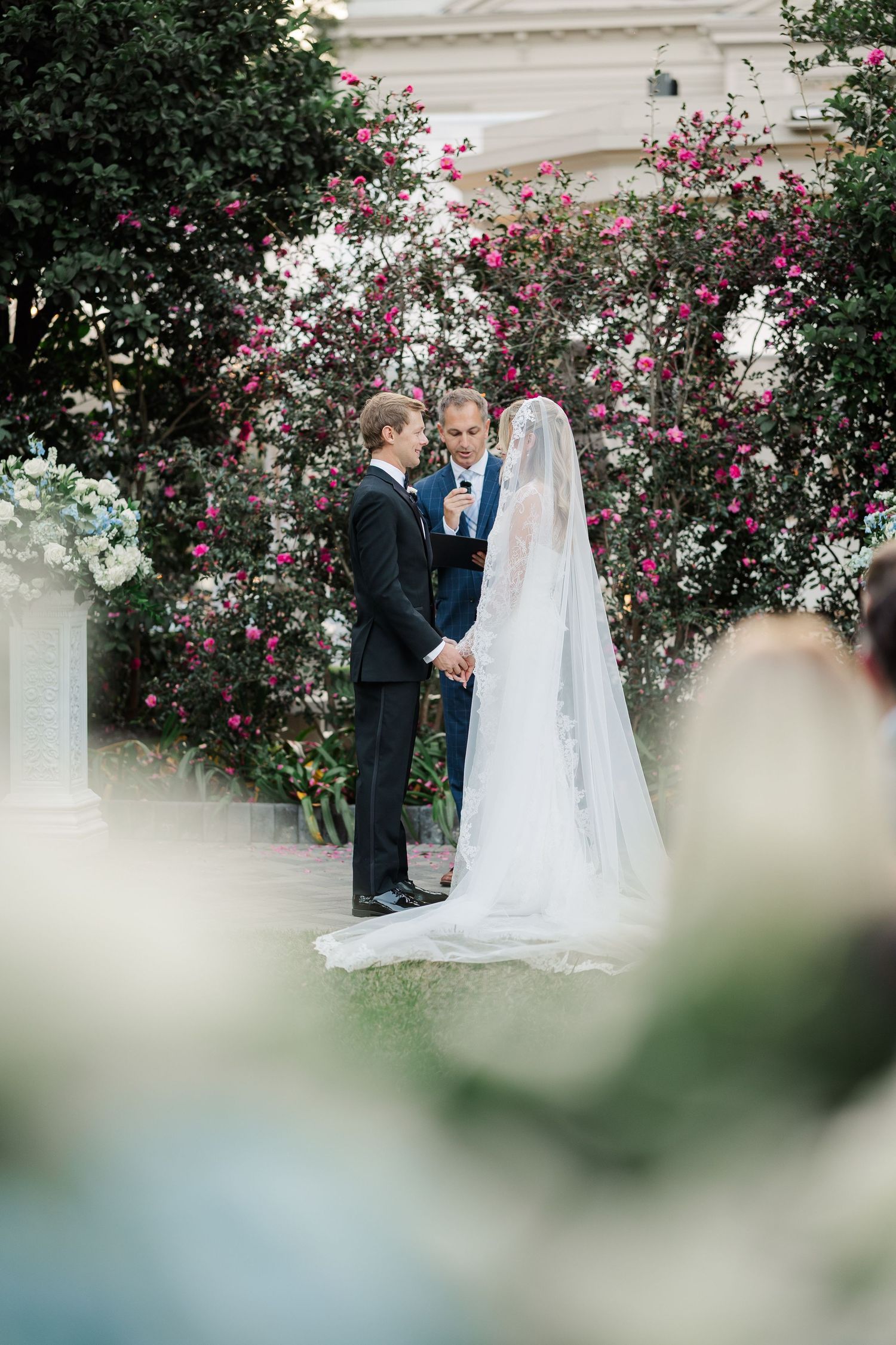 A wedding ceremony takes place in a garden setting with blooming rose bushes and classic architecture.