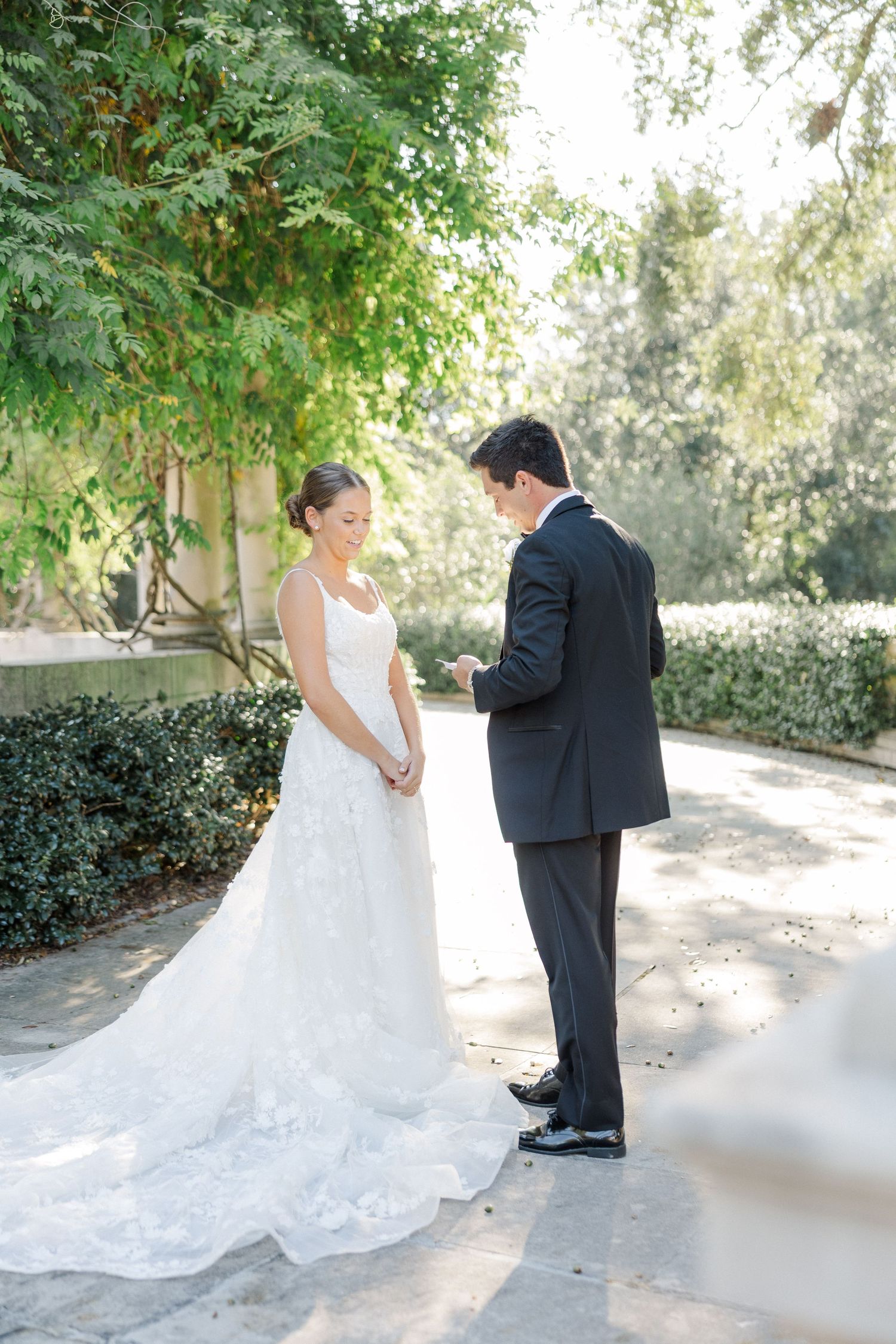 A romantic wedding first look moment in a garden setting with sunlit trees and a flowing white lace gown.