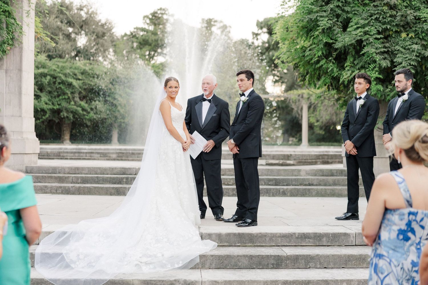 Wedding ceremony takes place outdoors with stone architecture and greenery in the background.