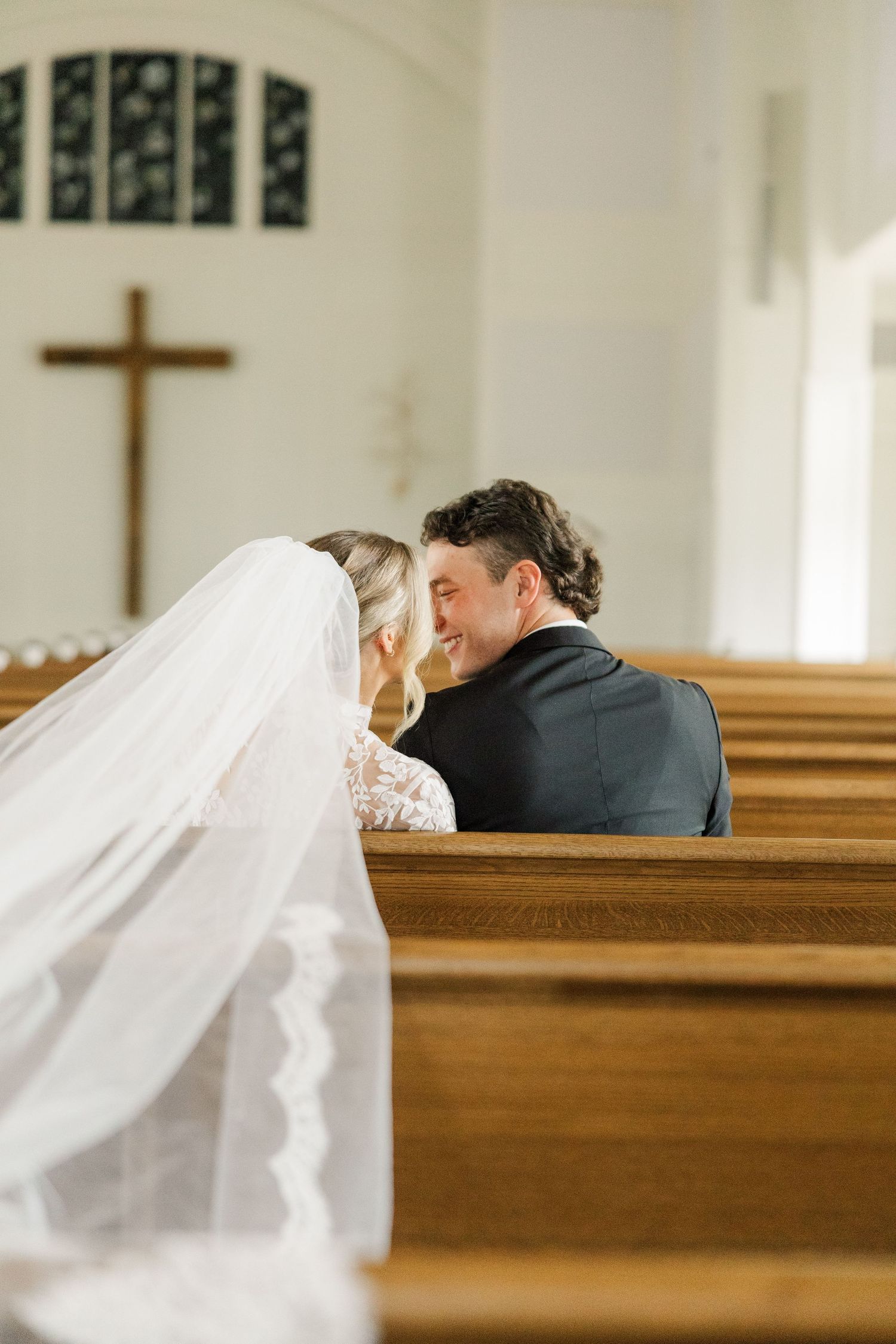 A couple shares an intimate moment in wooden church pews with a cross visible in background.