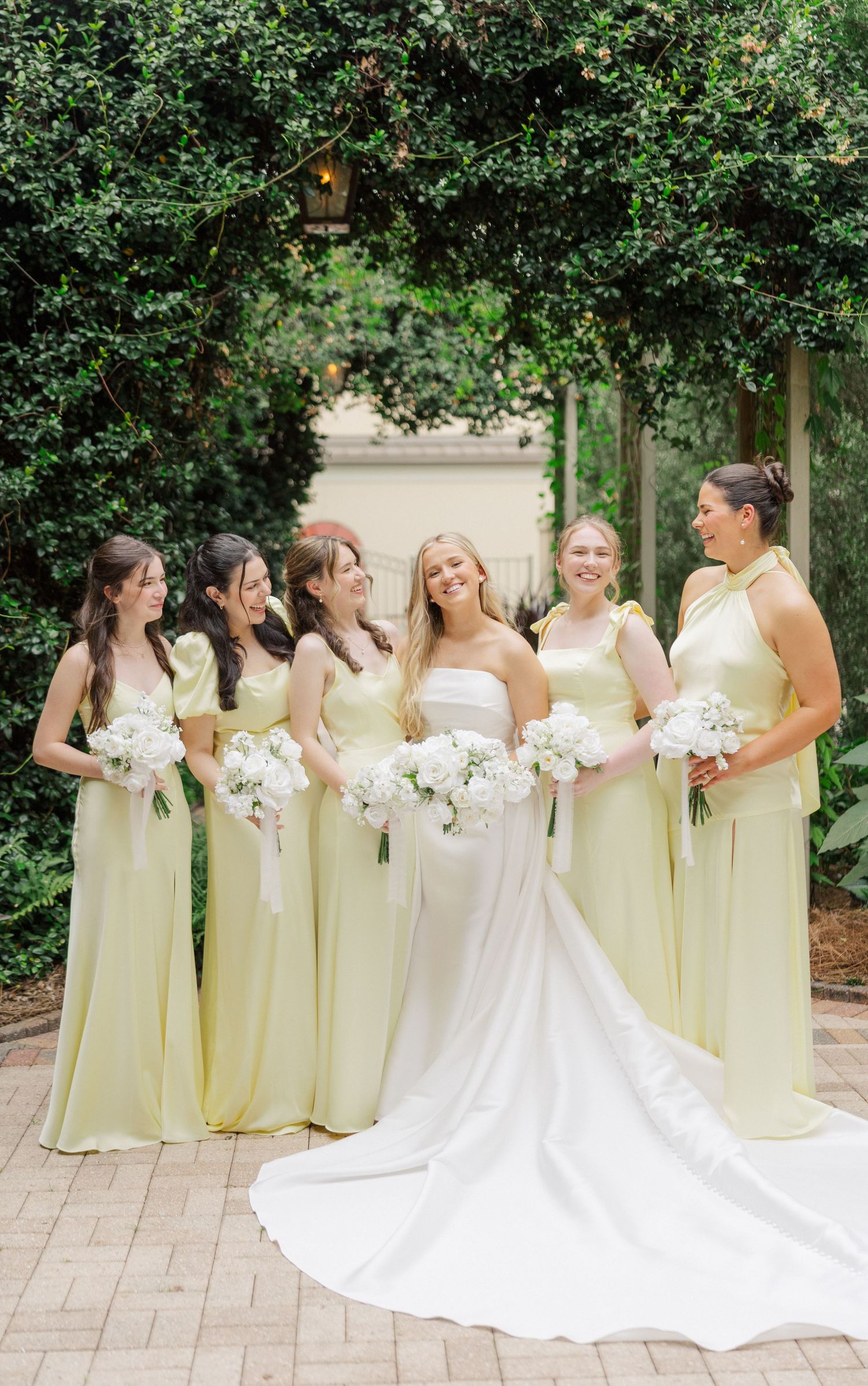 Wedding party poses outdoors with bridesmaids in pale yellow dresses holding white bouquets.