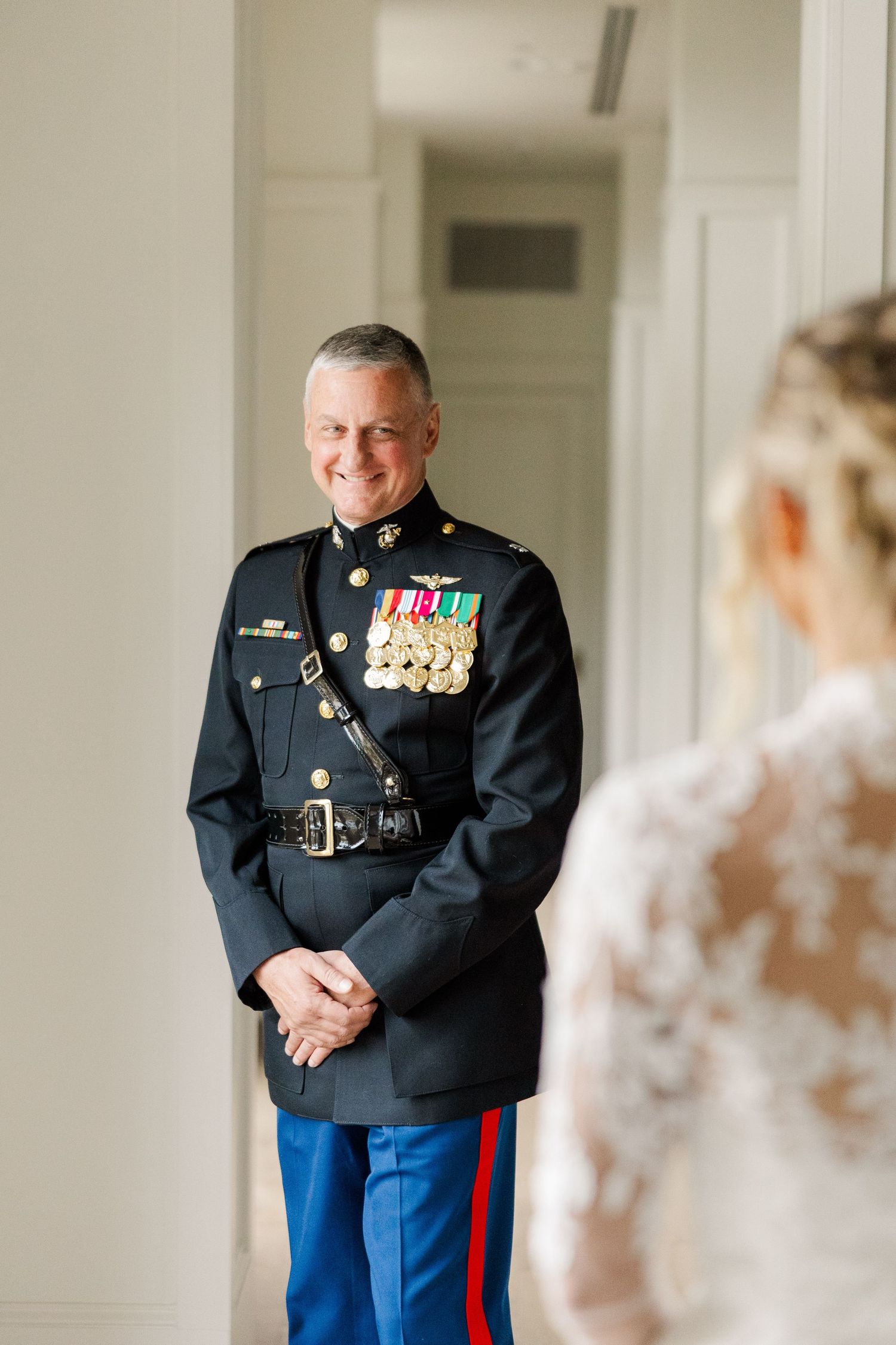 A Marine in dress uniform smiles during a wedding first look moment in an elegant white hallway.