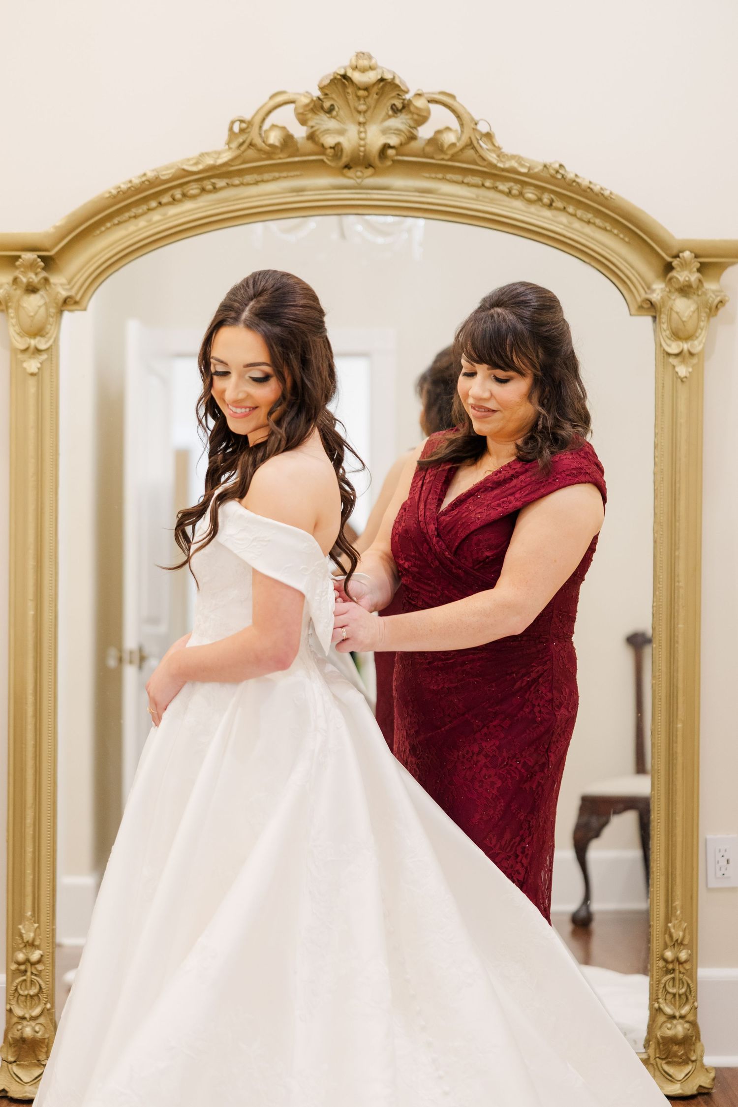 A woman in burgundy helps adjust a white wedding dress in front of ornate gold-framed mirrors.