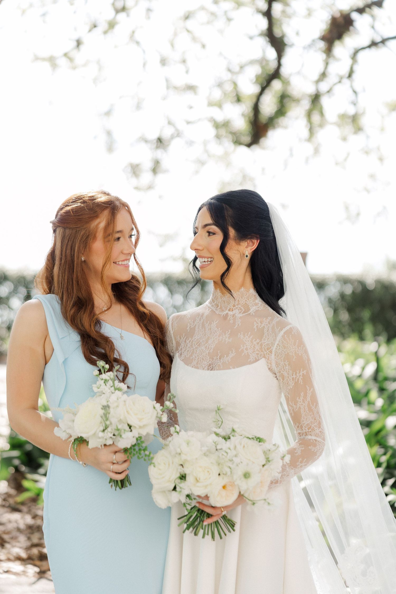 Bride in lace wedding gown smiles with bridesmaid in light blue dress outdoors.