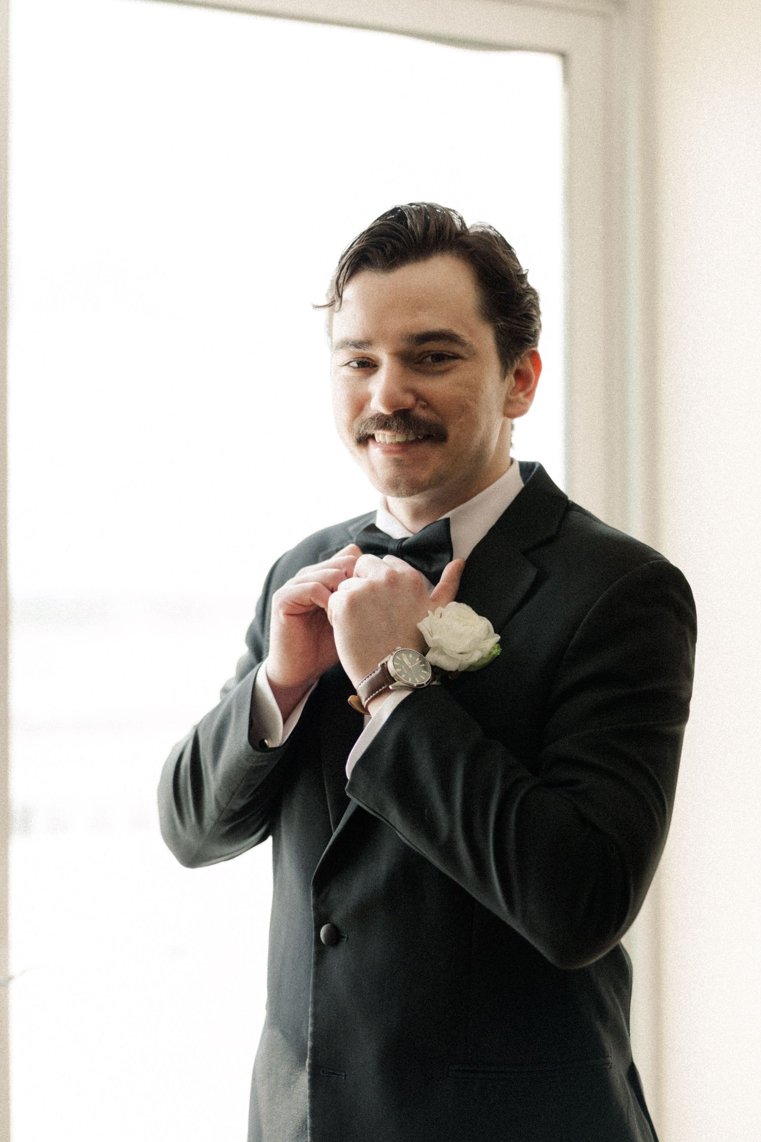 Smiling groom in a dark suit with bow tie and white boutonniere adjusting his cufflinks by a bright window.