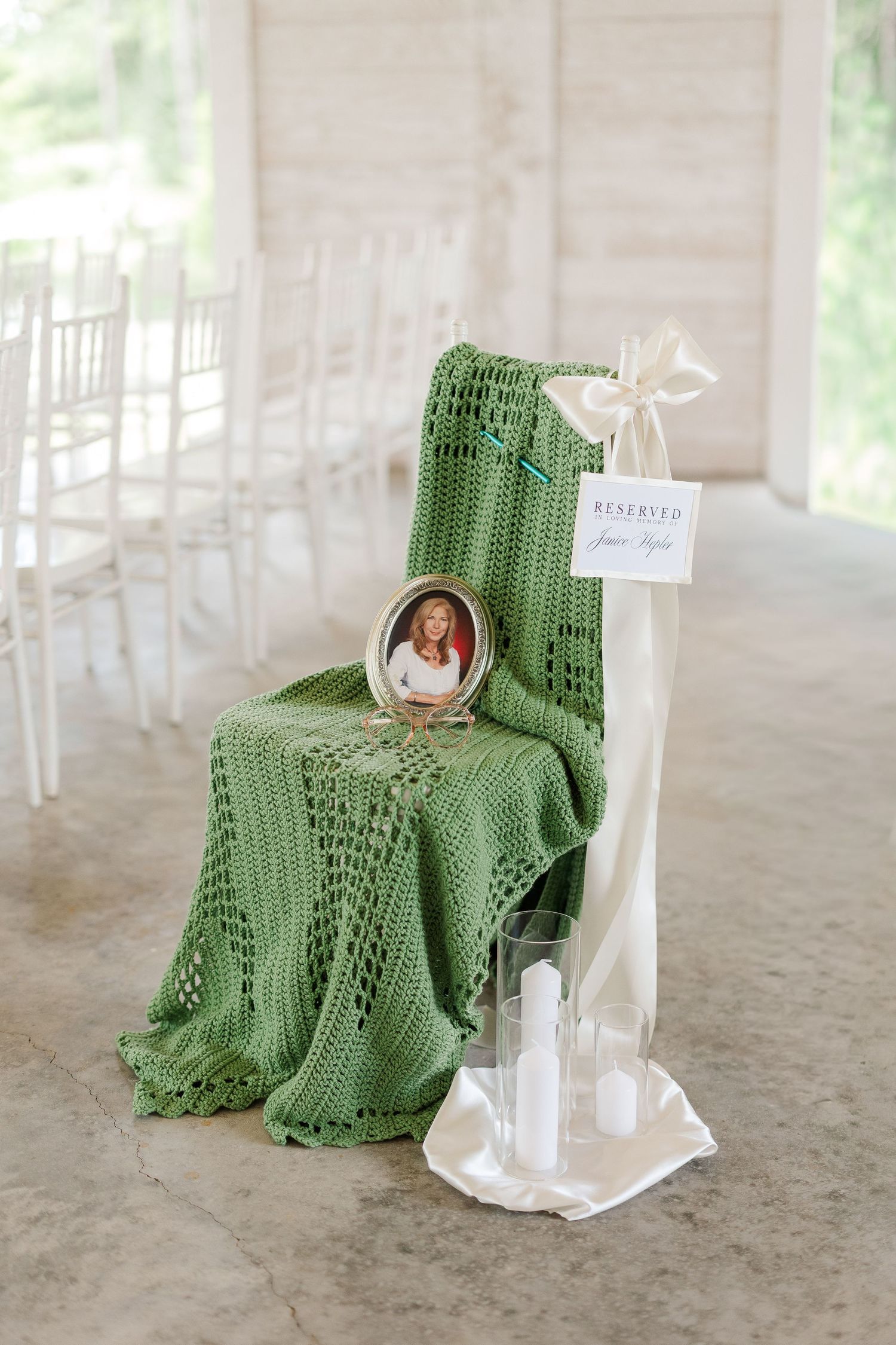 Reserved memorial chair draped with green crochet blanket, framed photo, white candles, and ribbon at wedding ceremony.