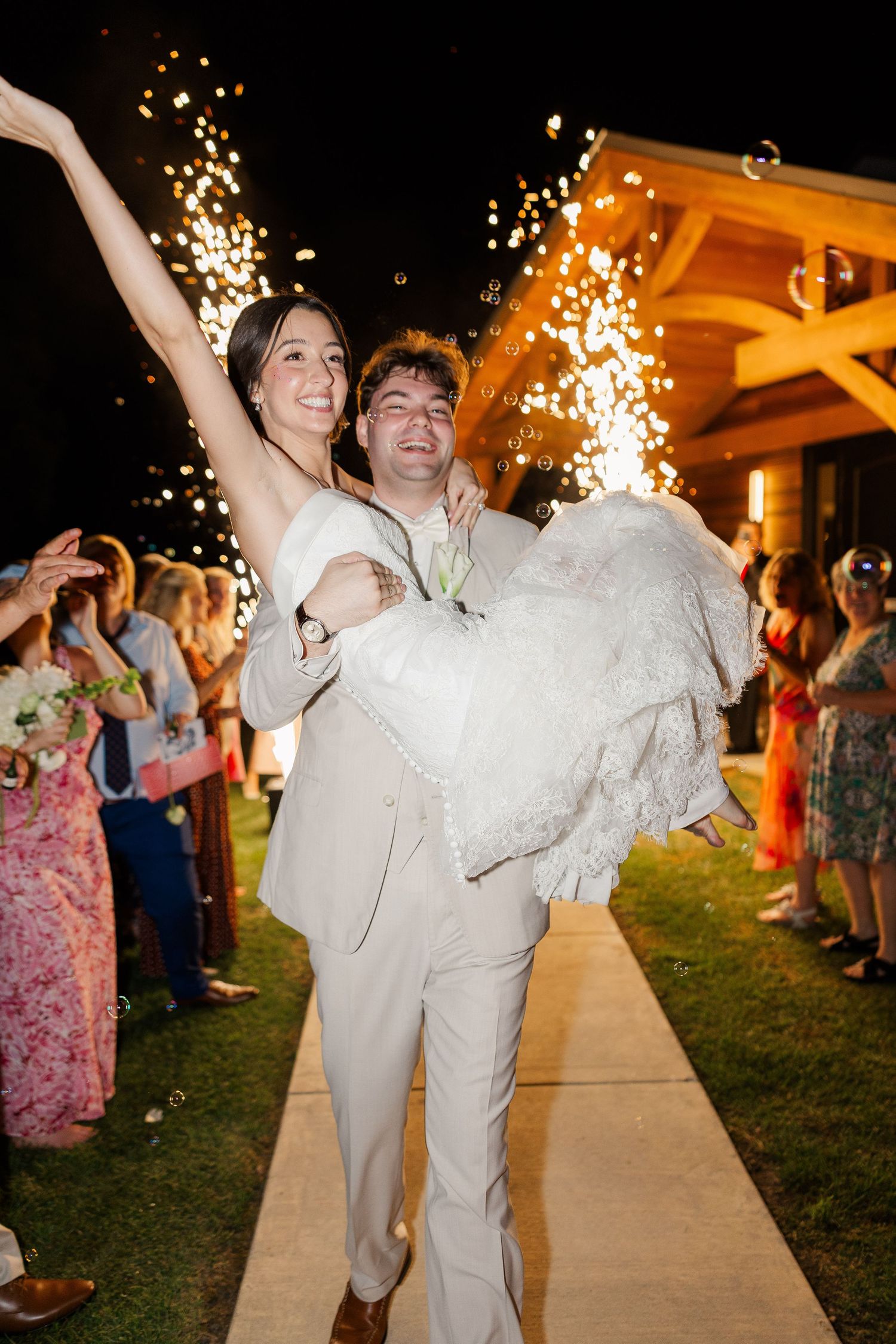 Groom carries beaming bride during sparkler grand exit at night, guests lining the pathway celebrating.