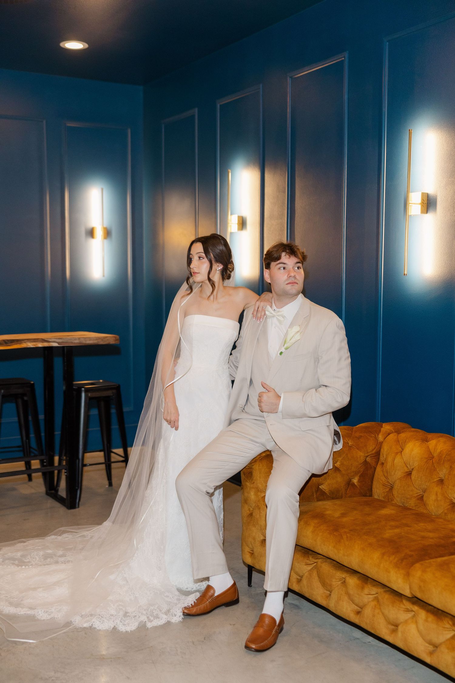 Bride and groom pose in elegant blue room with gold accents, groom seated on velvet sofa, bride standing.